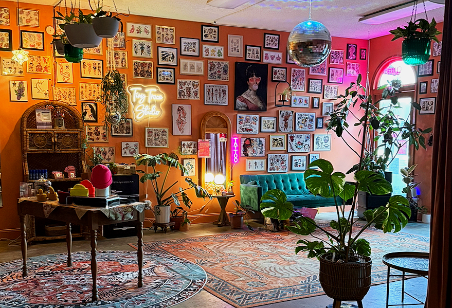 Interior of a colorful tattoo studio with framed art on the walls, a neon sign reading "Tiny Trace Studio," a green velvet sofa, a wooden table with art supplies, and various potted plants, including a Monstera.
