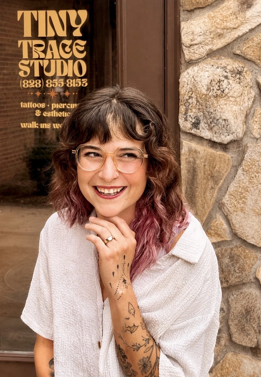 A woman with glasses and shoulder-length wavy hair, dyed pink at the ends, standing outside a tattoo studio with a stone wall and door in the background. She is smiling, with her hand resting on her chin, showing tattoos on her forearm.