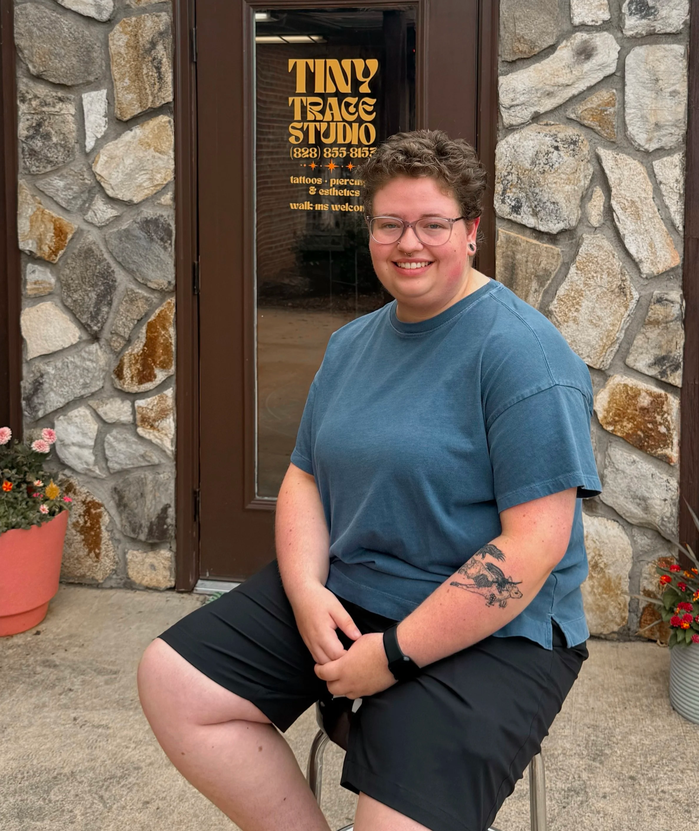 A smiling person with short curly hair, glasses, and a septum piercing, wearing a blue t-shirt and black shorts, sitting outside in front of a stone wall and door with a sign for Tiny Trace Studio, which offers tattoos and piercings.