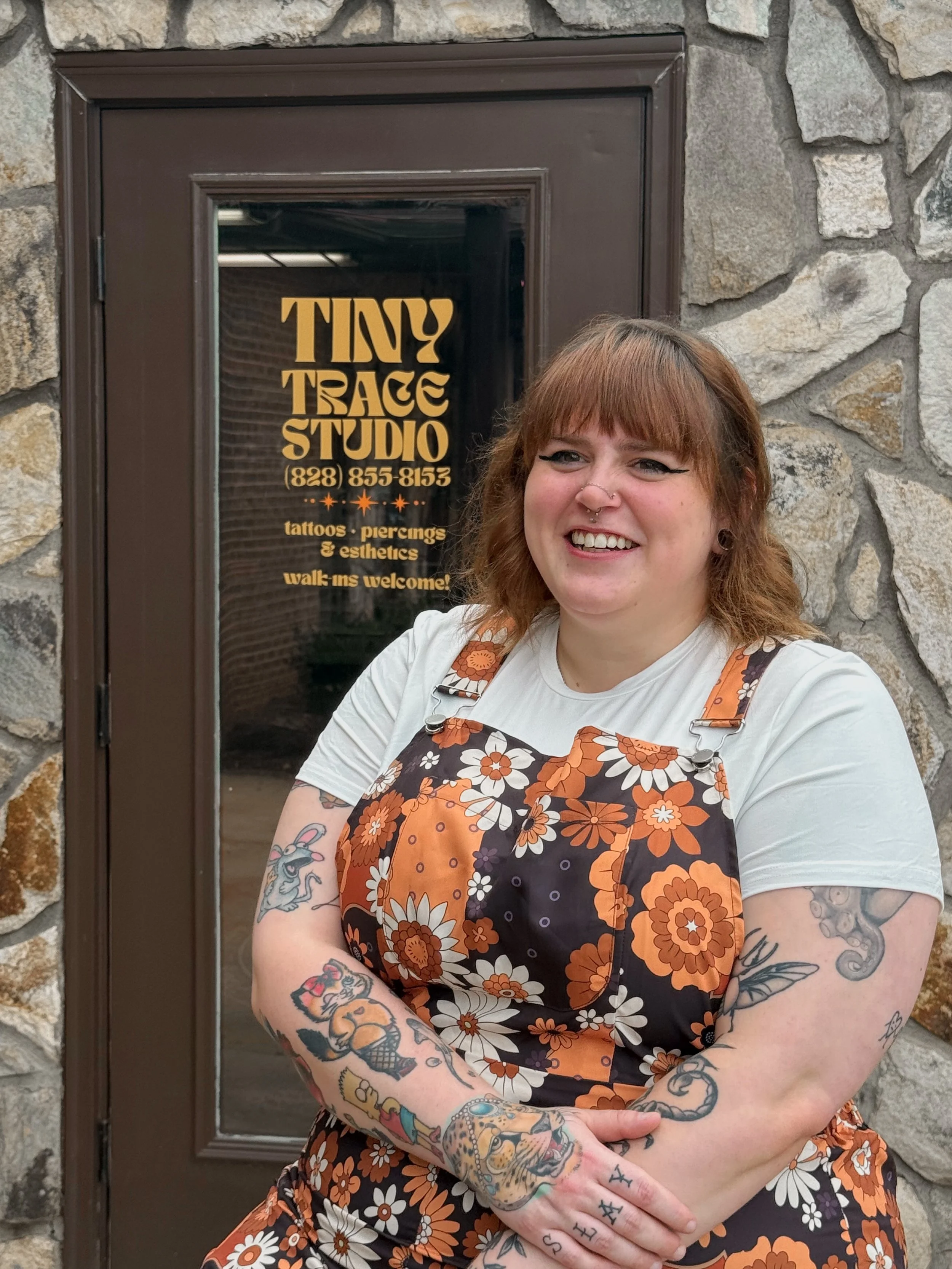 A person with red hair and tattoos smiling in front of a door with a sign for a tattoo studio, wearing a floral apron over a white shirt.