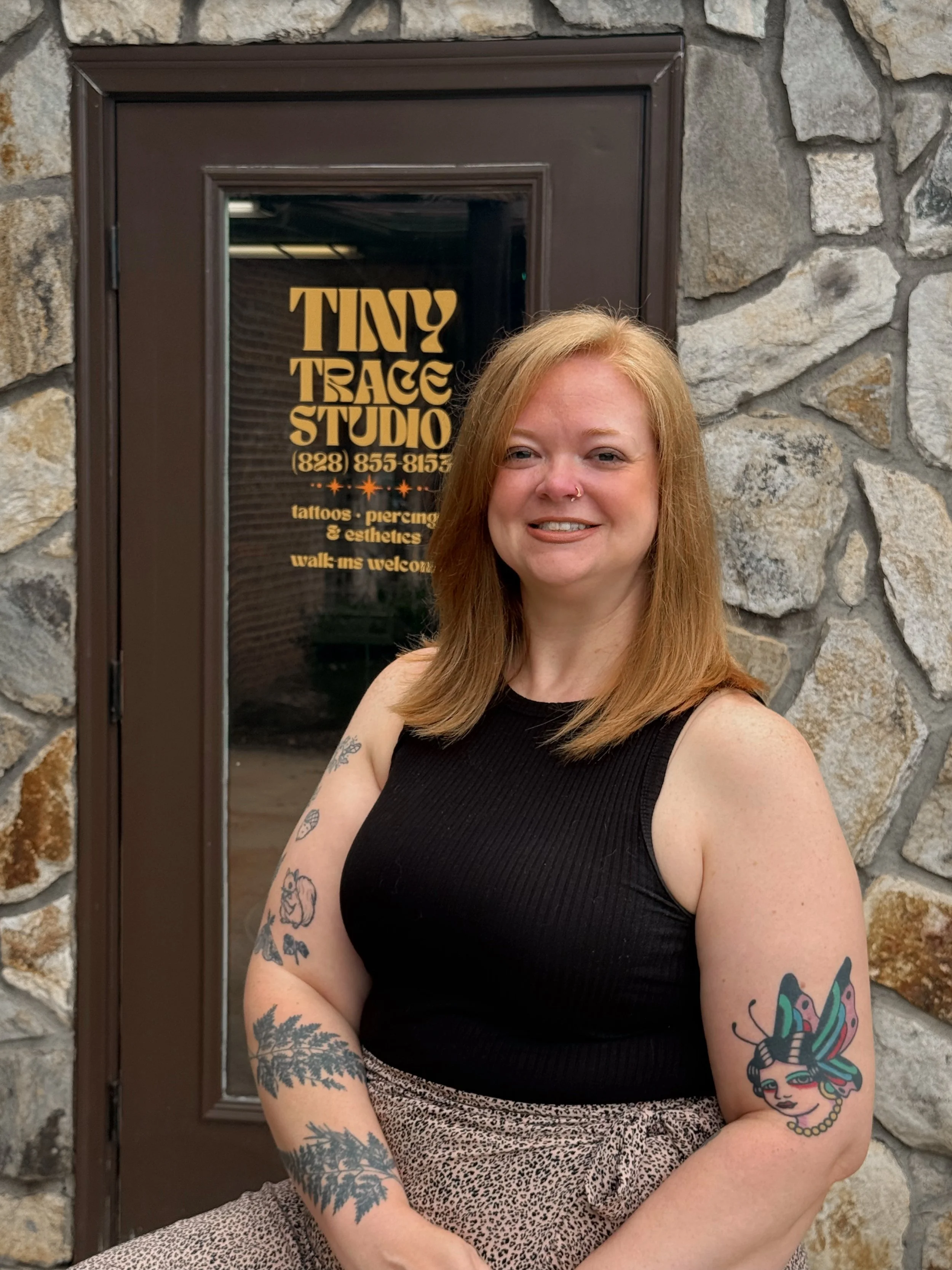 A woman with red hair and tattoos on her arms standing in front of a stone wall and a door with a sign for a tattoo studio.