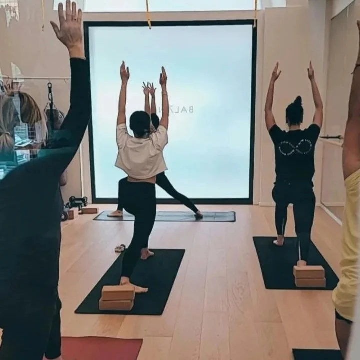 People participating in a yoga class practicing tree pose with blocks, facing a screen displaying the word 'BALANCE' in a studio.