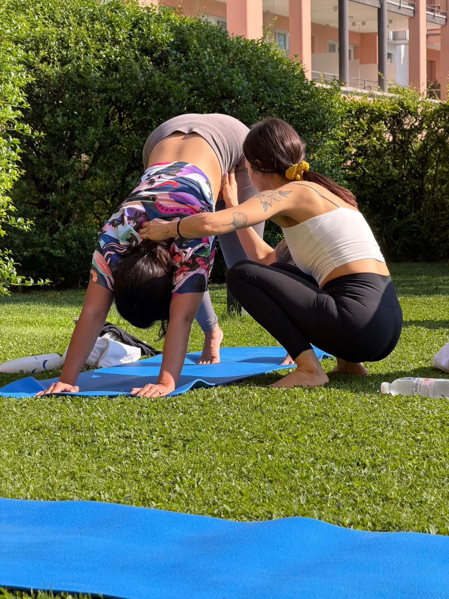 Two women practicing yoga outdoors, one in a downward dog pose and the other assisting, on a grassy area with bushes and an apartment building in the background.