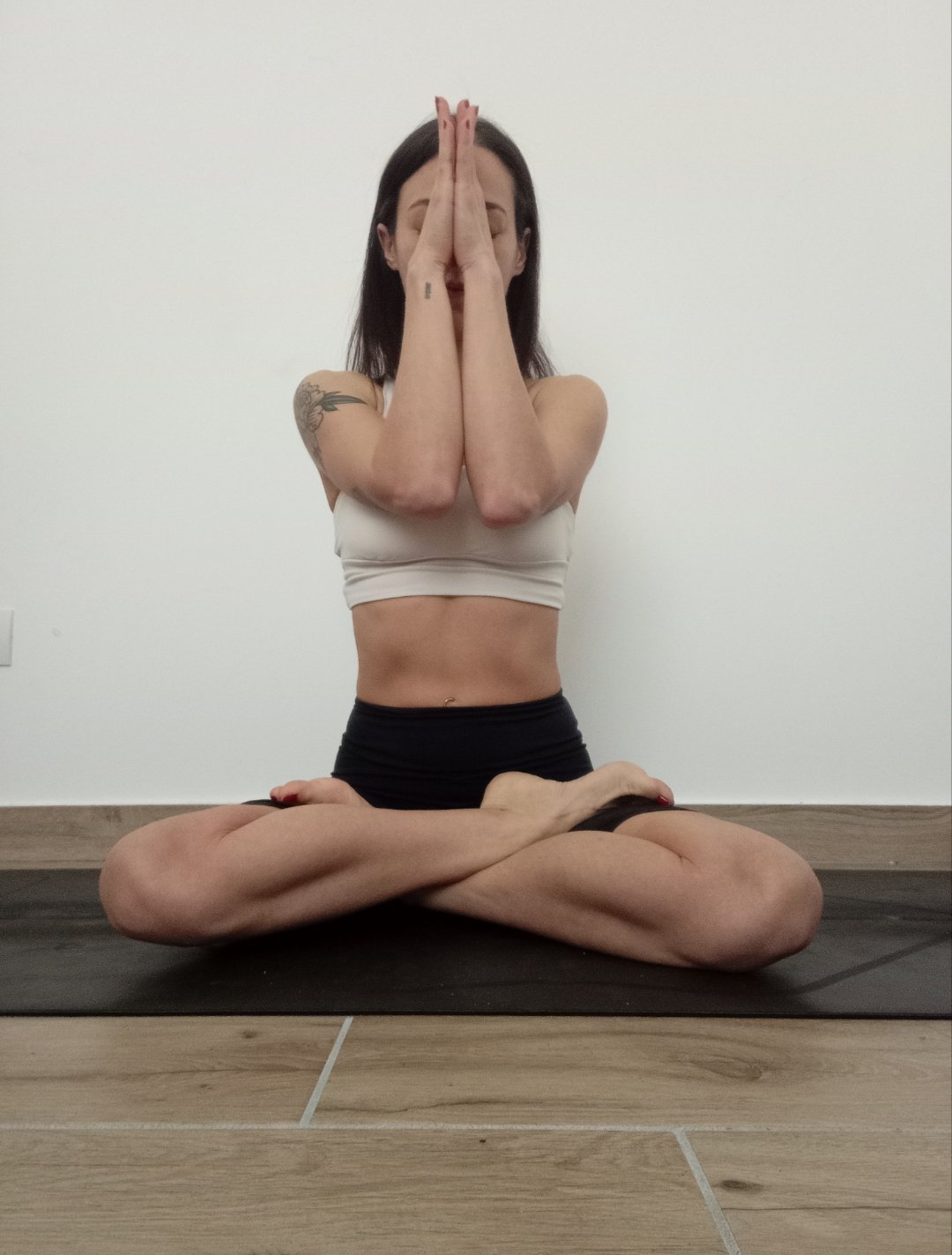 Woman practicing yoga in a seated cross-legged pose, with hands pressed together in prayer position, in front of a plain white wall.