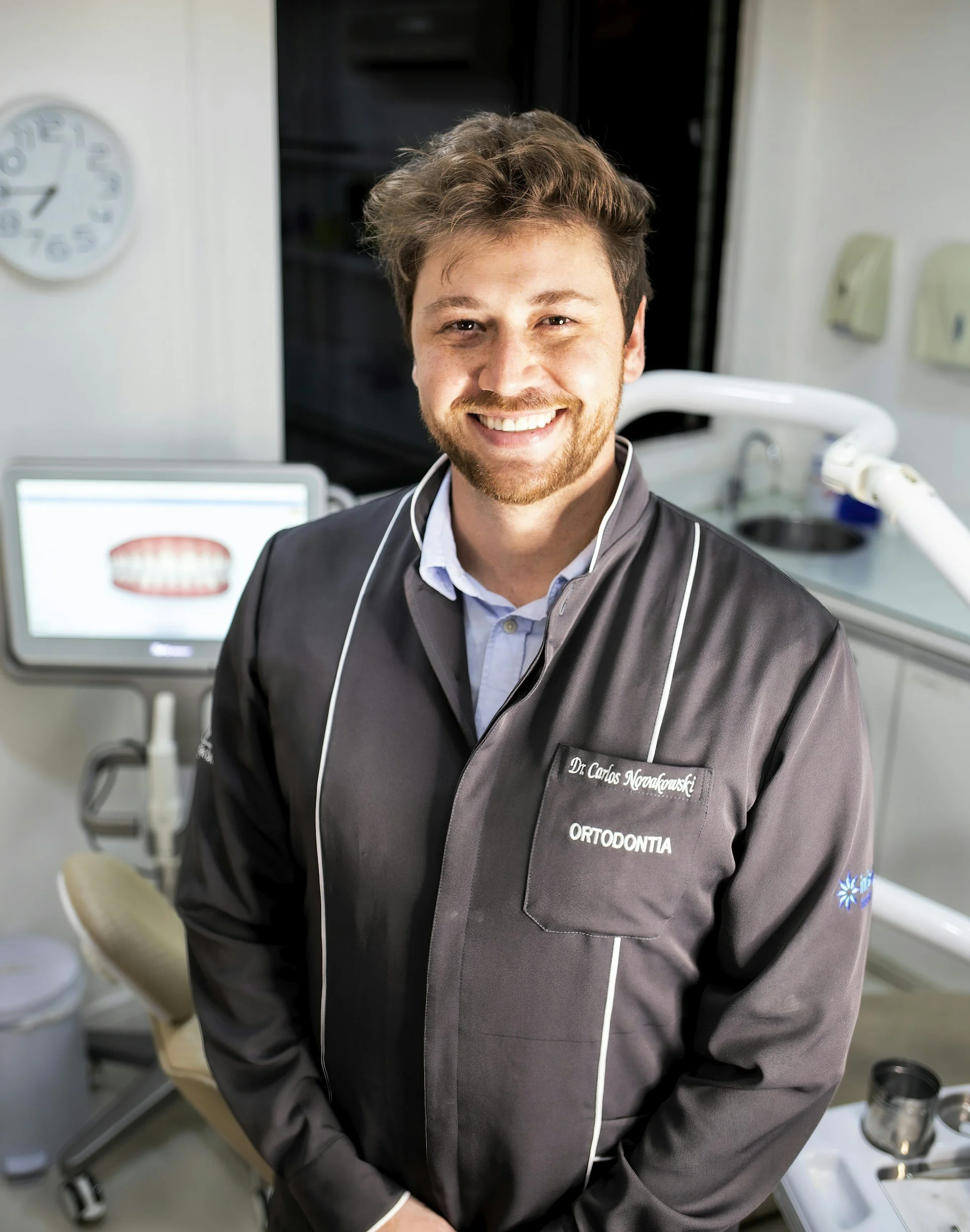 Smile of a young man in a medical uniform inside a dental clinic, standing in front of dental equipment and a computer screen.