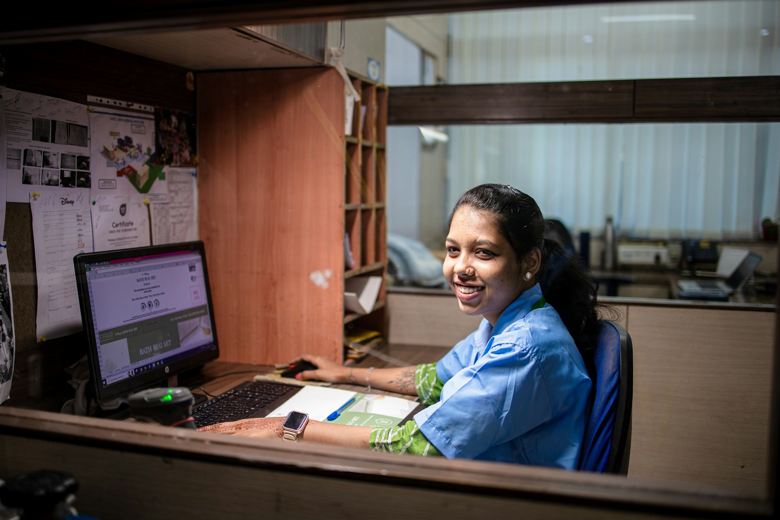 A woman sitting at a desk in an office, smiling, wearing a blue shirt and a smartwatch, with a computer, papers, and a bulletin board in the background.