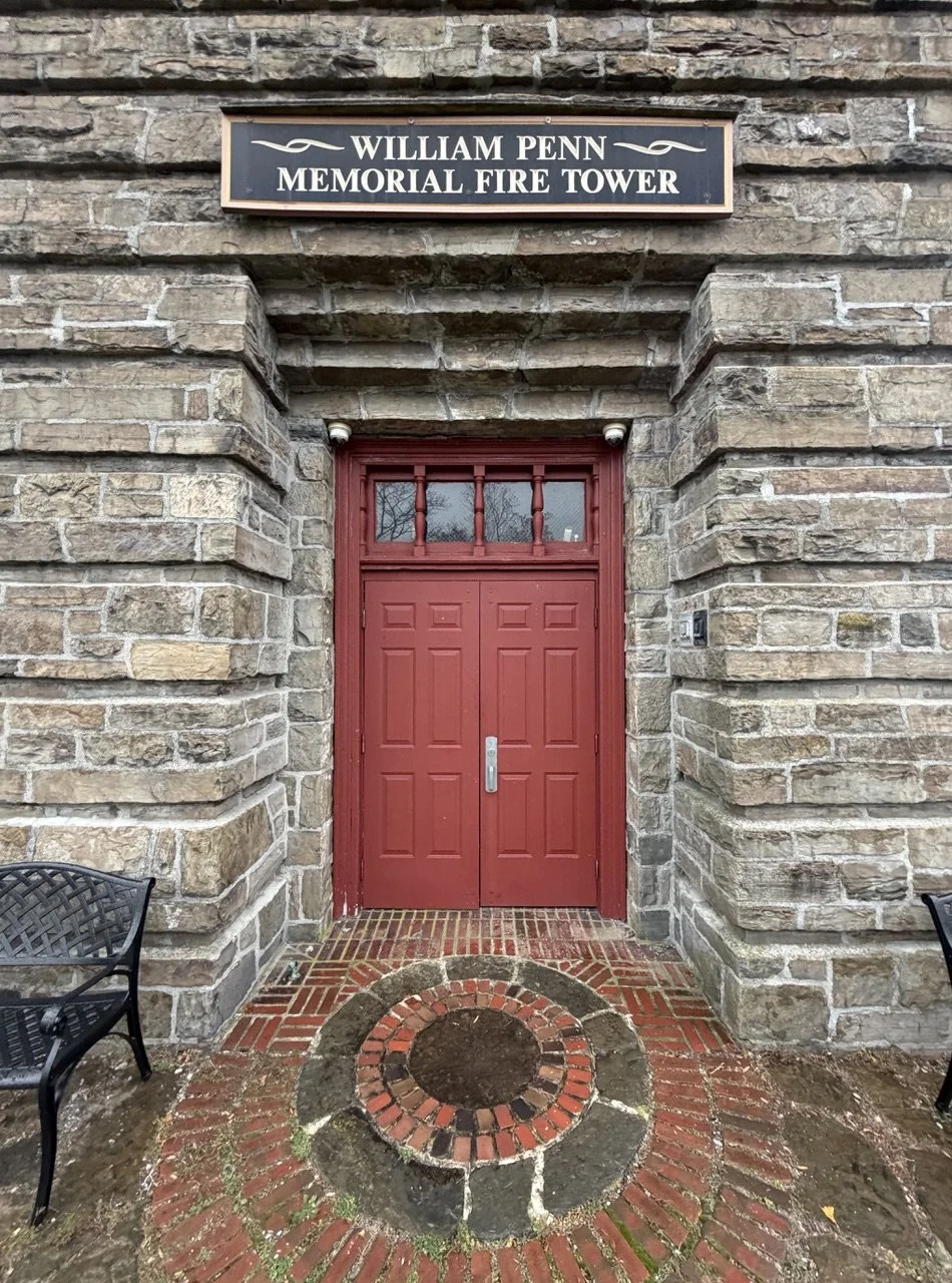 Brick building with a red door, two small benches on either side, and a sign above that reads "William Penn Memorial Fire Tower."