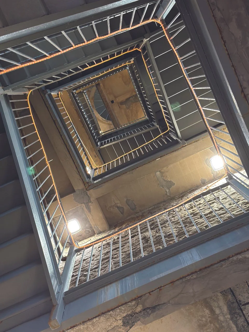 Interior view looking up the stairwell of a multi-story building, showing metal stair railings with string lights, concrete walls, and a brick floor at the bottom.