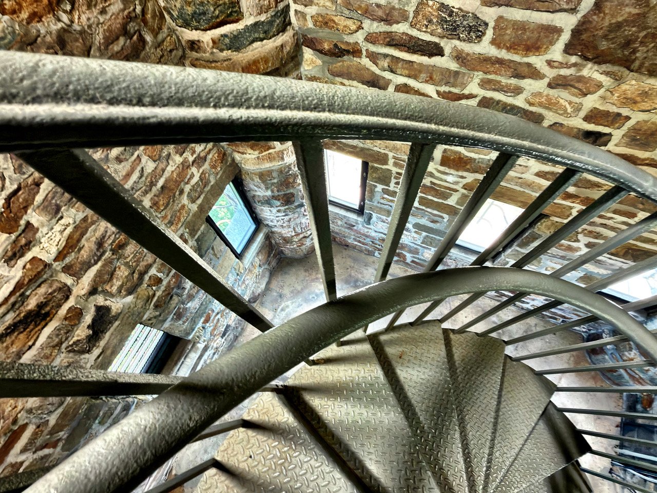 Spiral staircase with metal steps and railing inside a building with brick walls and small rectangular windows.