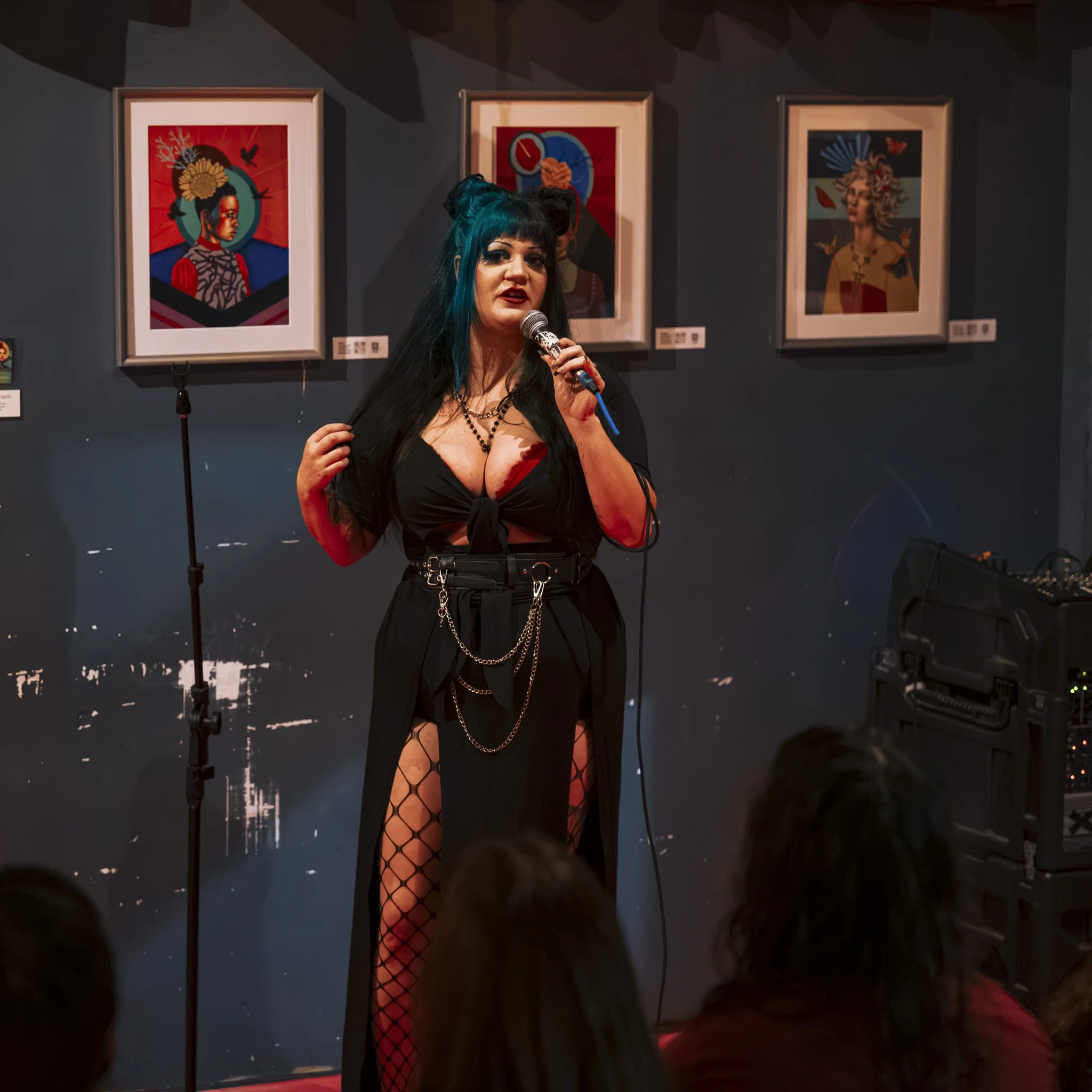 A woman with black and blue hair wearing fishnet stockings and black clothing sings into a microphone at an art gallery or exhibition, with framed artwork hanging on a dark-colored wall behind her.