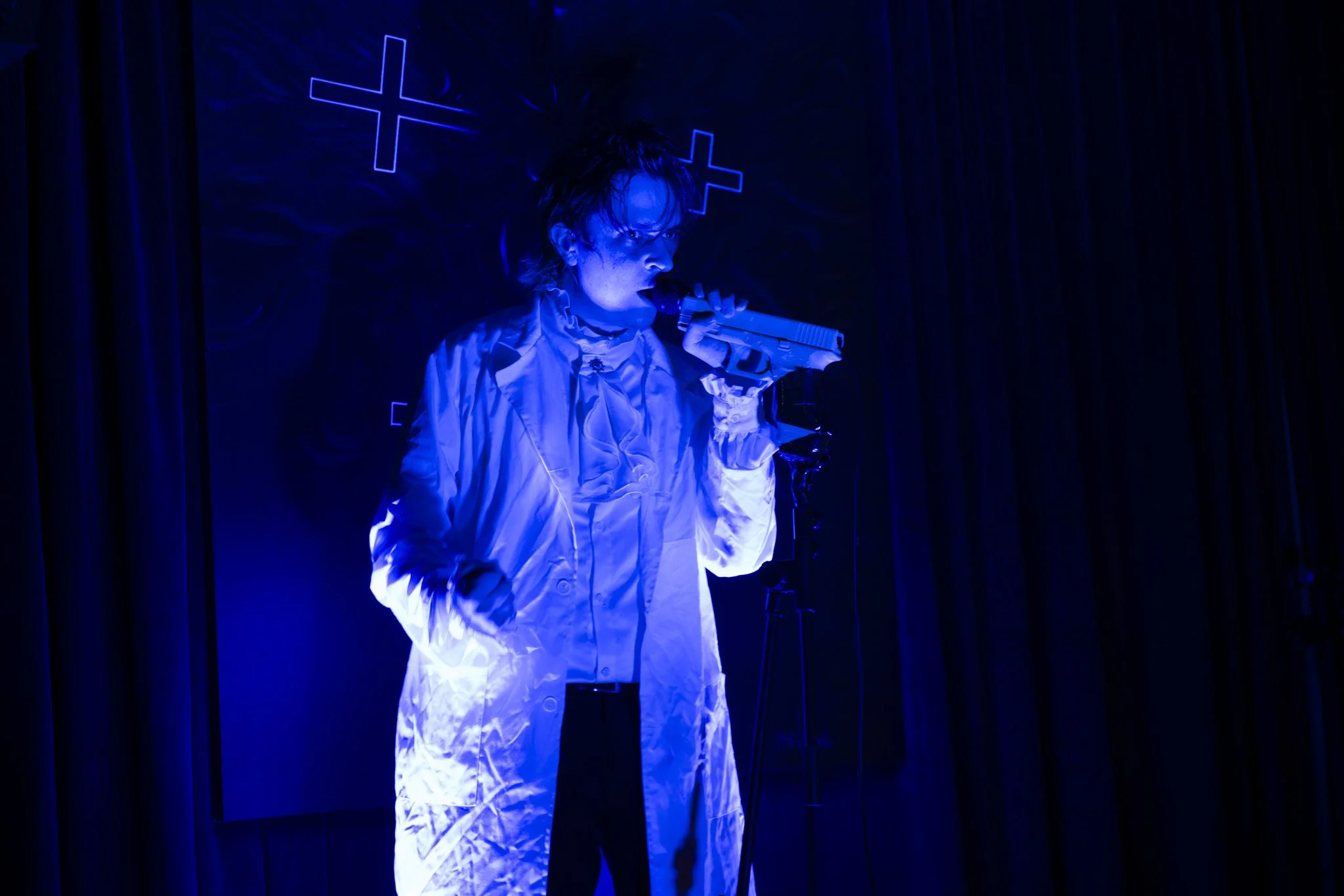 A person with wet hair wearing a white jacket, holding a microphone to their mouth, standing on a stage with blue lighting and crosses illuminated on a dark background.