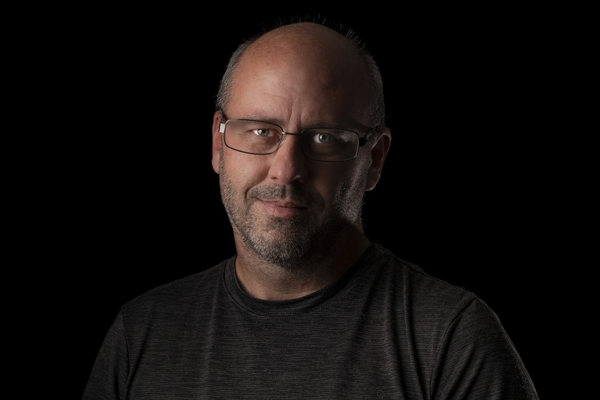 Close-up portrait of a middle-aged man with glasses, light beard, wearing a black shirt, against a black background.