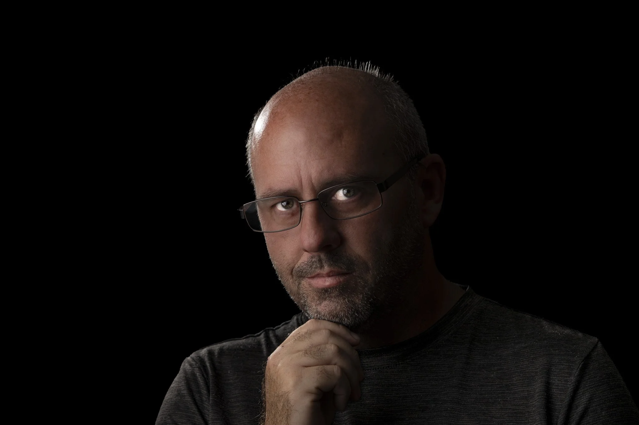 Close-up portrait of a middle-aged man with glasses, short gray hair, and a gray beard, resting his chin on his hand, set against a plain black background.