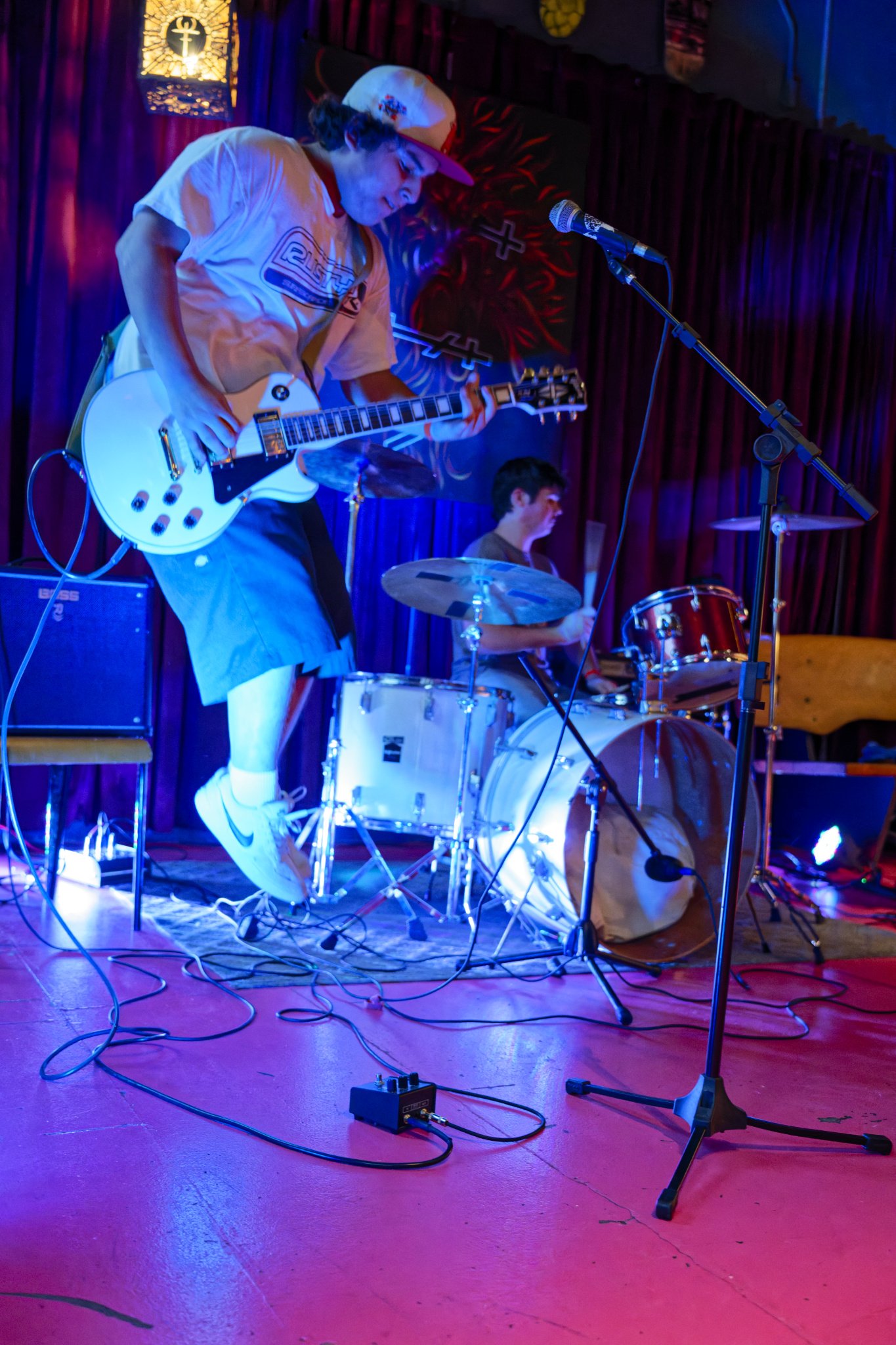 A live music performance with a guitarist and drummer on stage, illuminated by colorful stage lighting, in a venue with a red floor and dark curtains.
