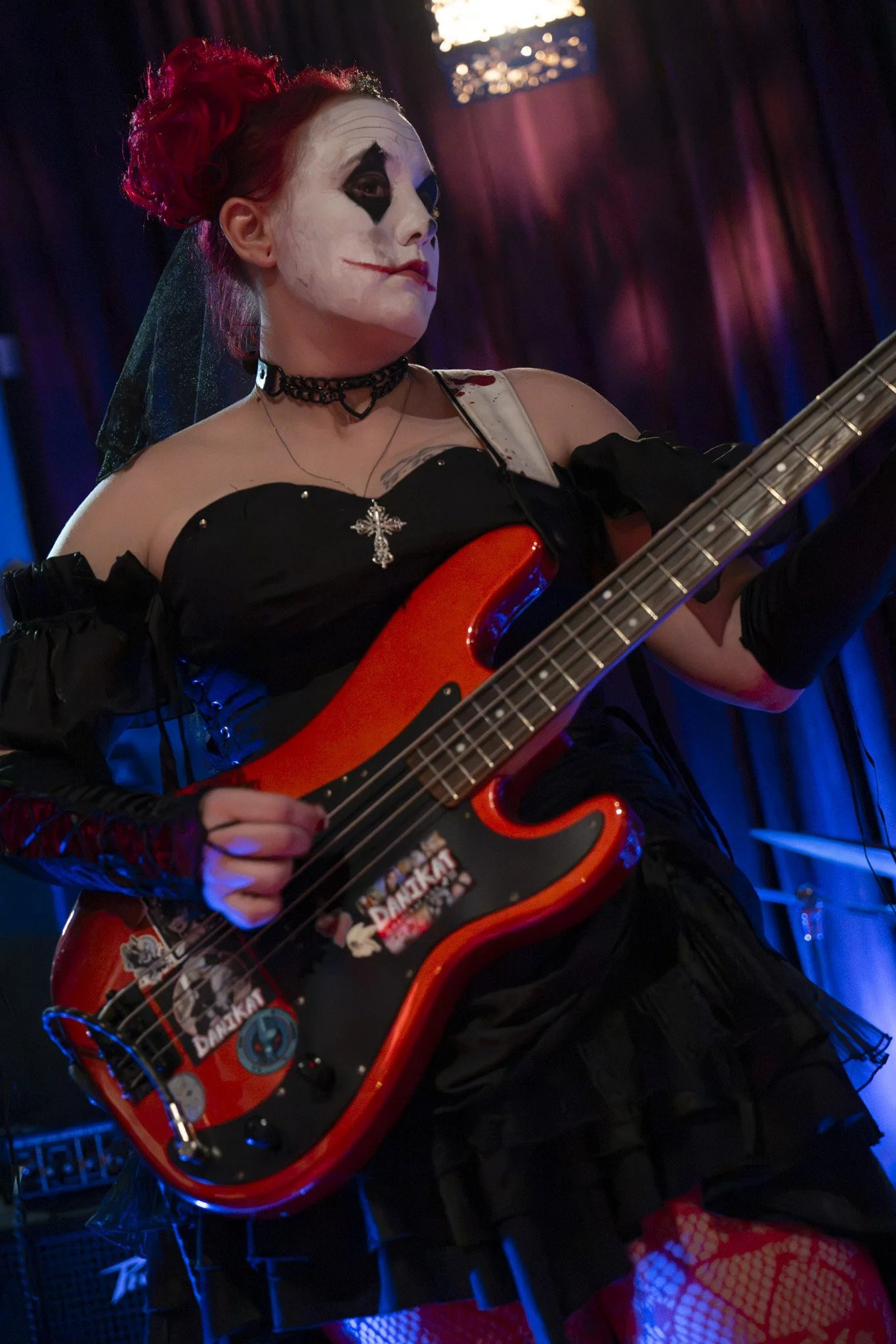 Person with face makeup resembling a clown, red hair, black choker, holding a red electric guitar decorated with various stickers, in a dimly lit room with dark curtains and red lighting.