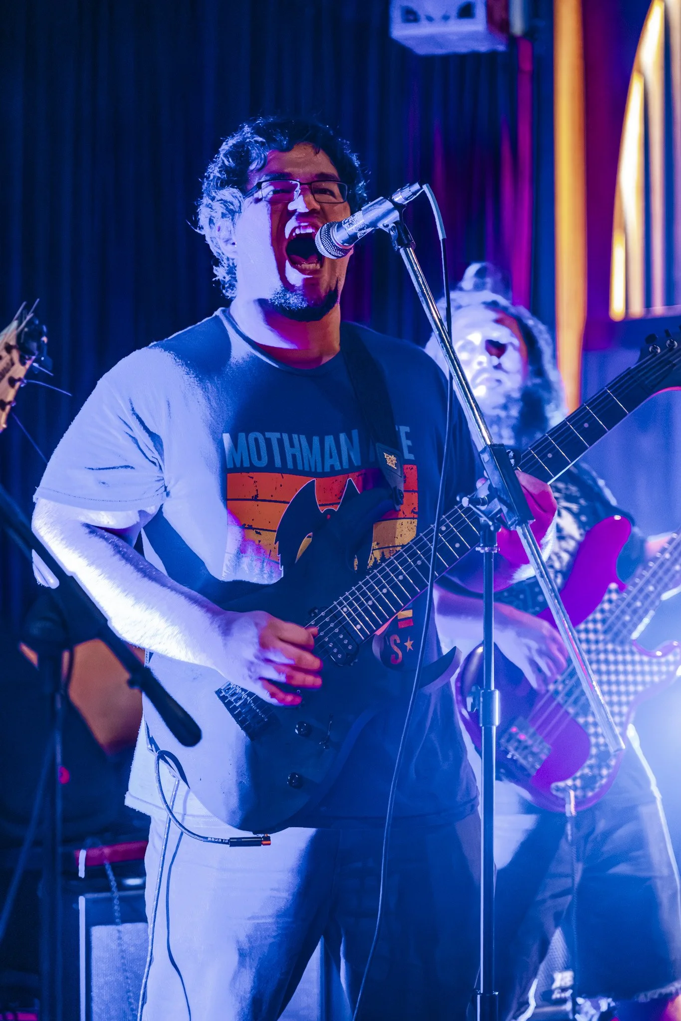 A man with glasses singing into a microphone while playing an electric guitar on stage with colorful lighting.