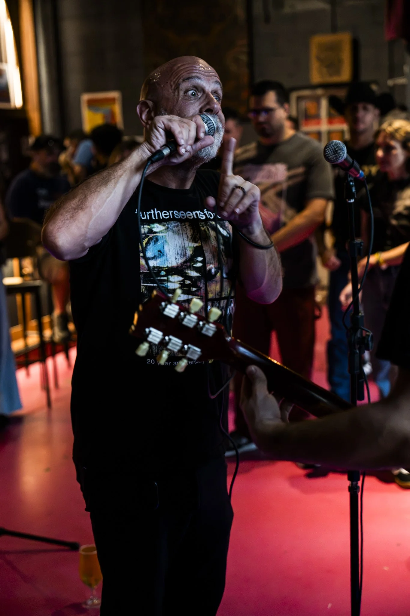 A man with a bald head and a beard is singing into a microphone while playing an electric guitar at an indoor event. He appears to be passionately performing, with a crowd and artwork displayed in the background.