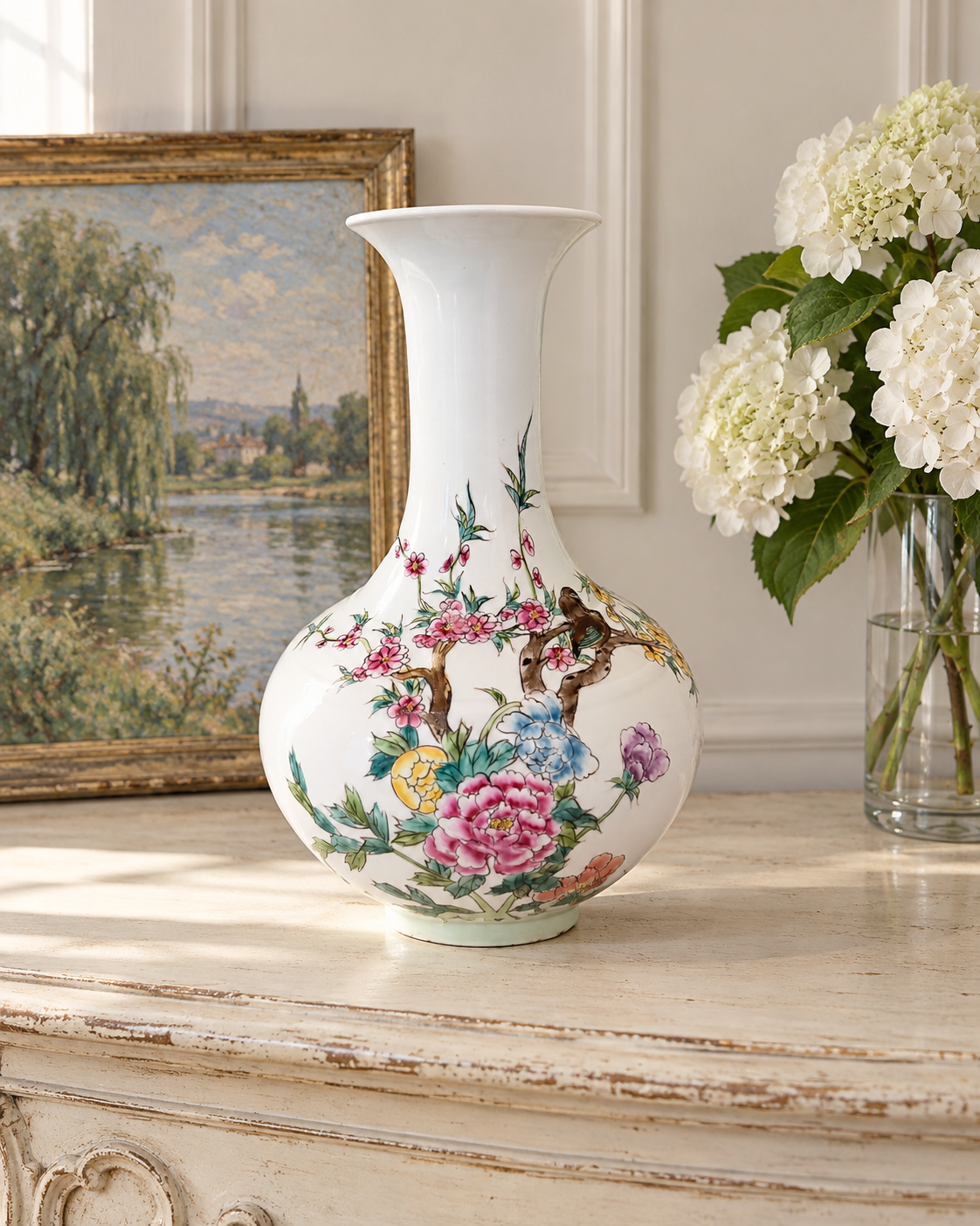 Famille rose style vase on an ivory French console beside framed landscape art and white hydrangeas.