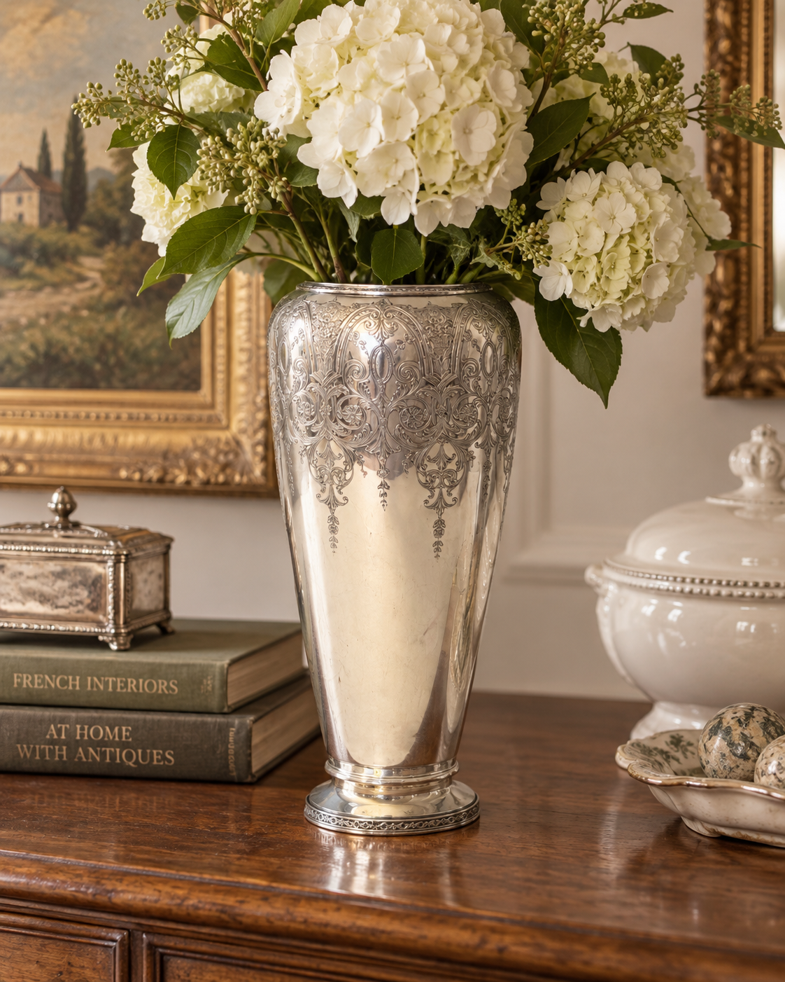 Antique silver-plated vase with pedestal base and ornate engraving, styled on an elegant chippy French sideboard.