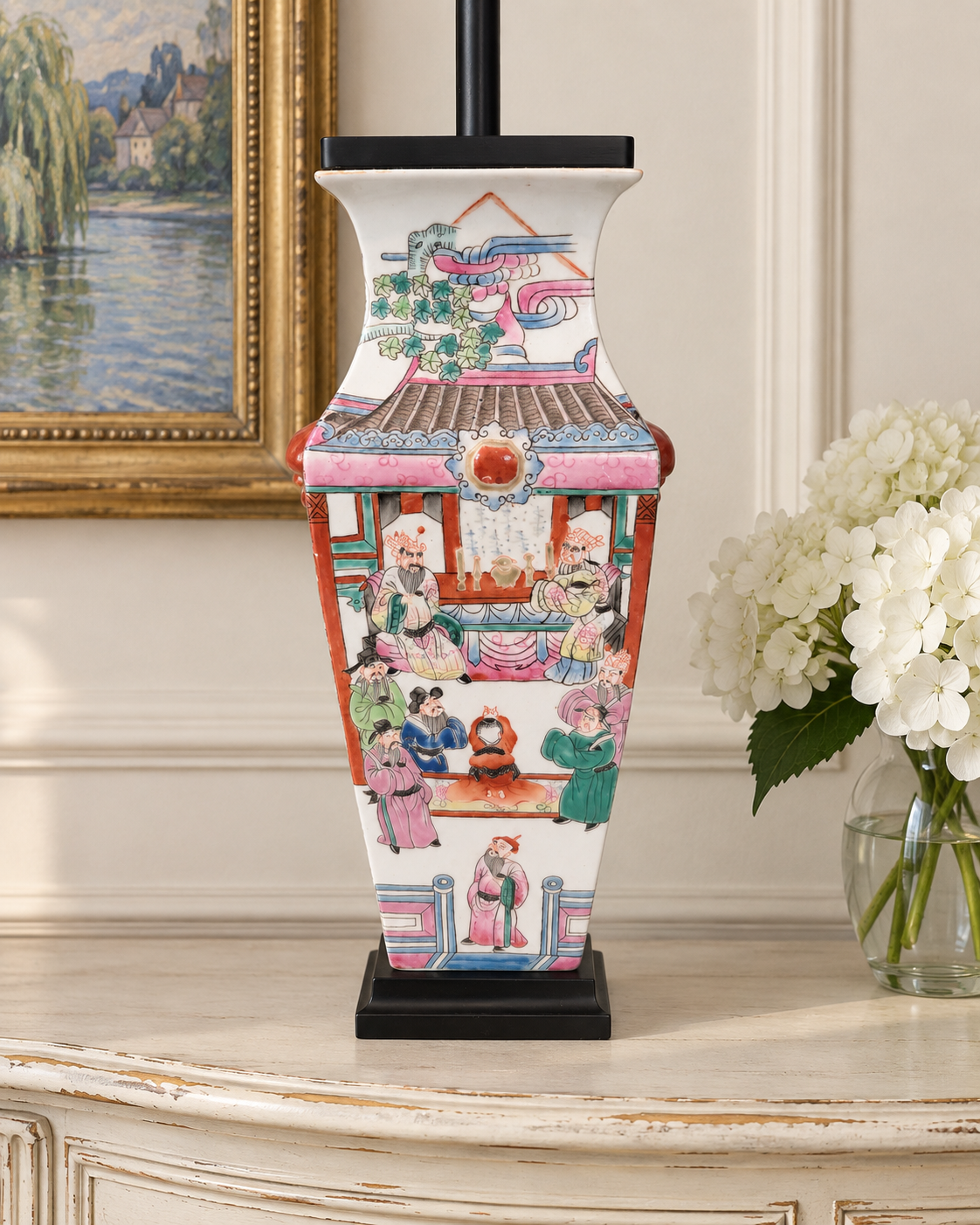 Reverse decoration of a porcelain lamp beside white hydrangeas and framed landscape art.
