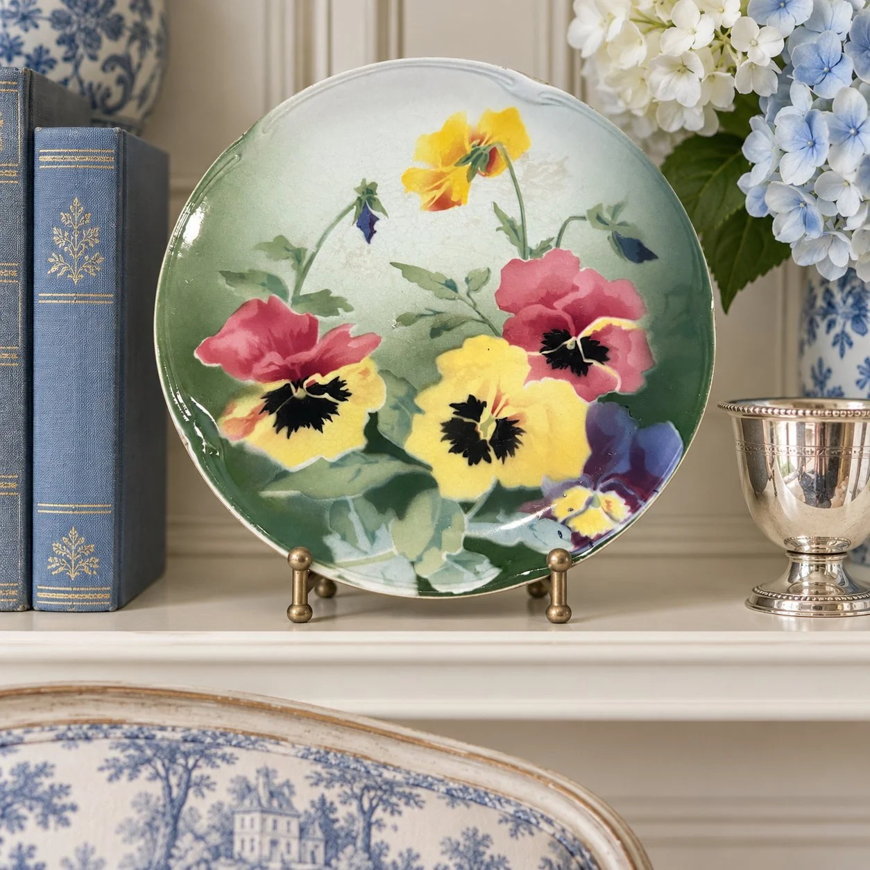 Lunéville pansy plate displayed on an ivory shelf with blue books and a silver accent.