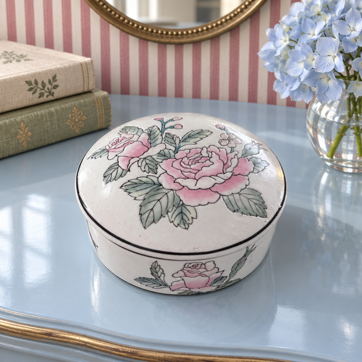 Pink floral porcelain box on a blue vanity with pale-blue hydrangeas and a brass-framed mirror.
