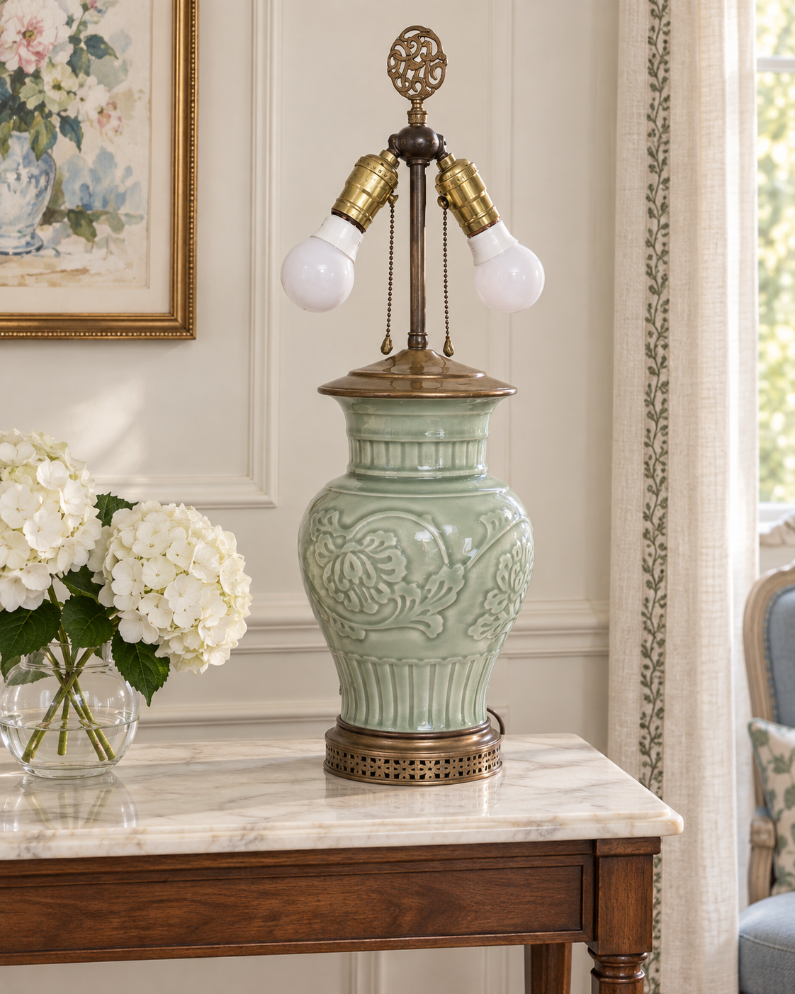 Celadon table lamp without a shade on a marble-topped console beside white hydrangeas.
