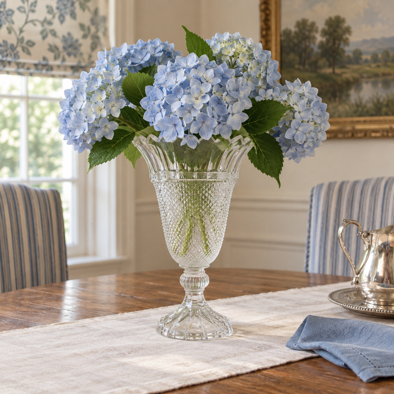 Clear pedestal vase with pale-blue hydrangeas on a linen runner in a dining room.