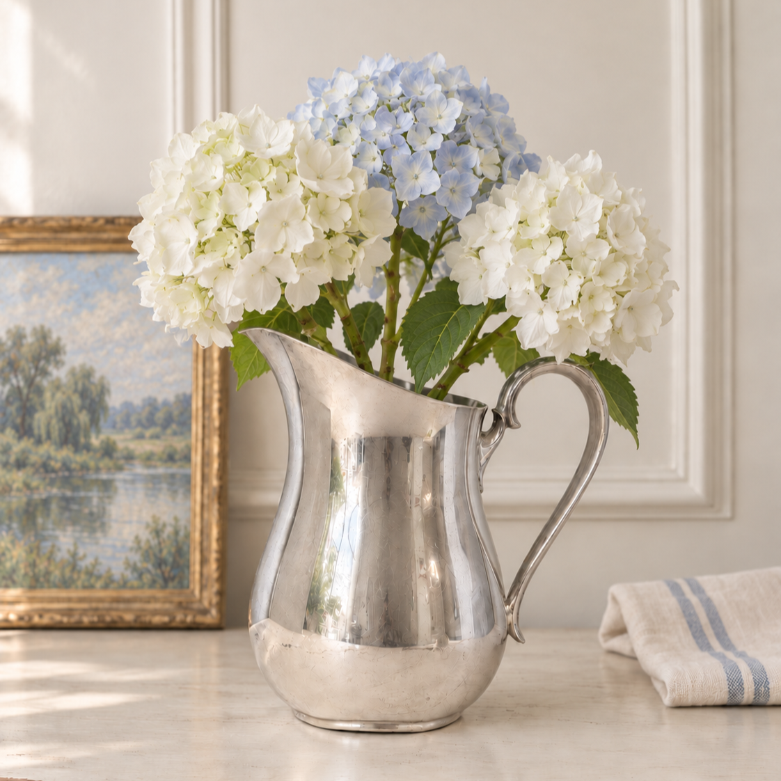 Silverplate pitcher with white and pale blue hydrangeas beside blue-striped linen in a softly lit French setting.