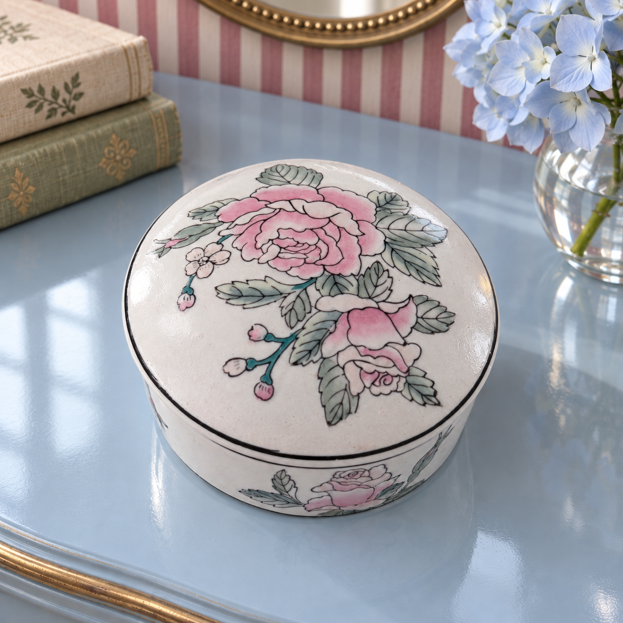 Pink floral porcelain box on a blue vanity with pale-blue hydrangeas and a brass-framed mirror.