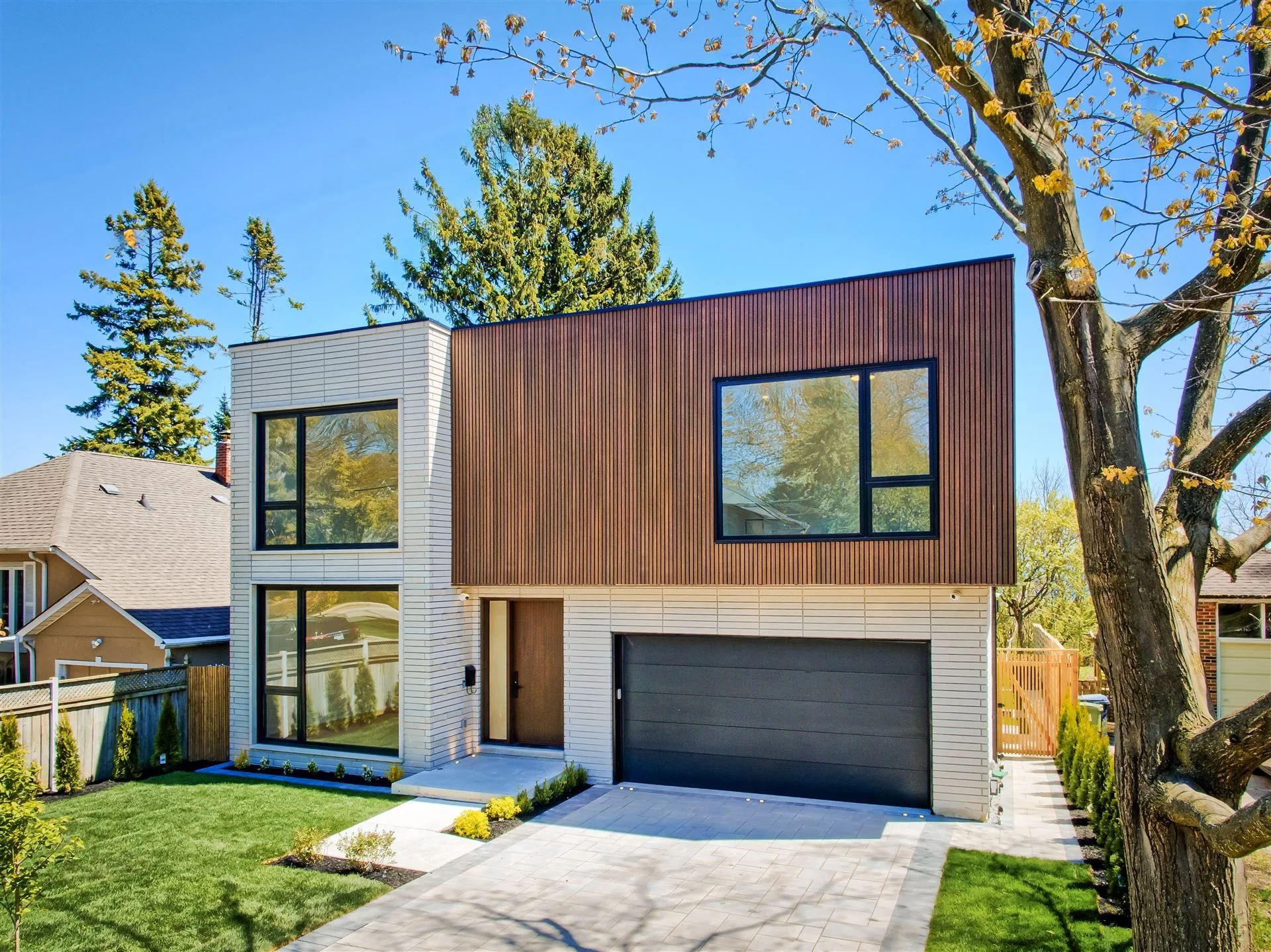 Modern two-story house with black and white exterior, large windows, and a garage, situated next to a tree and neighboring homes, with an ocean view in the distance.