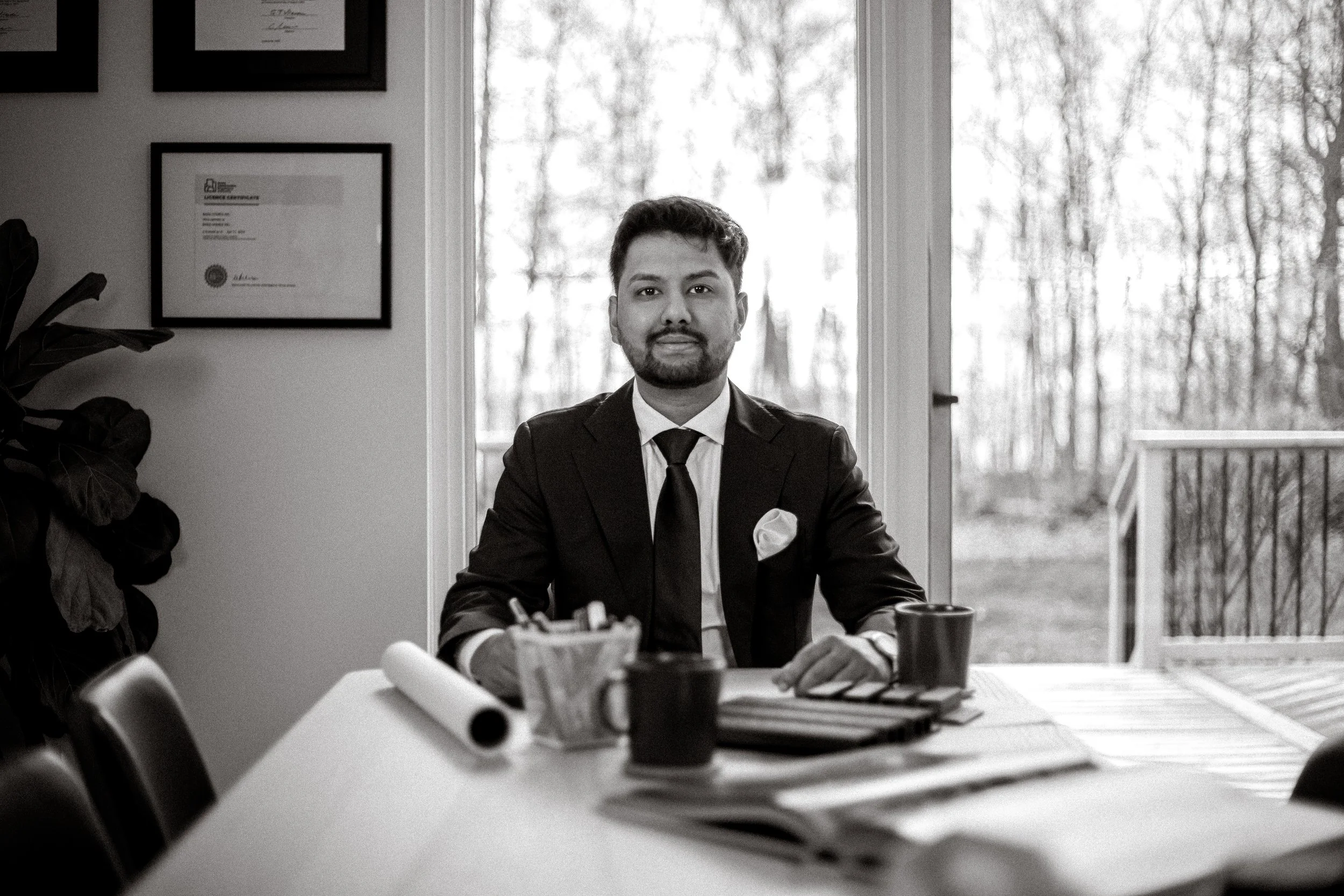 A man in a suit sitting at a conference table in a room with large windows showing trees outside. The table has papers, a coffee mug, and office supplies. There are framed certificates or awards on the wall behind him.