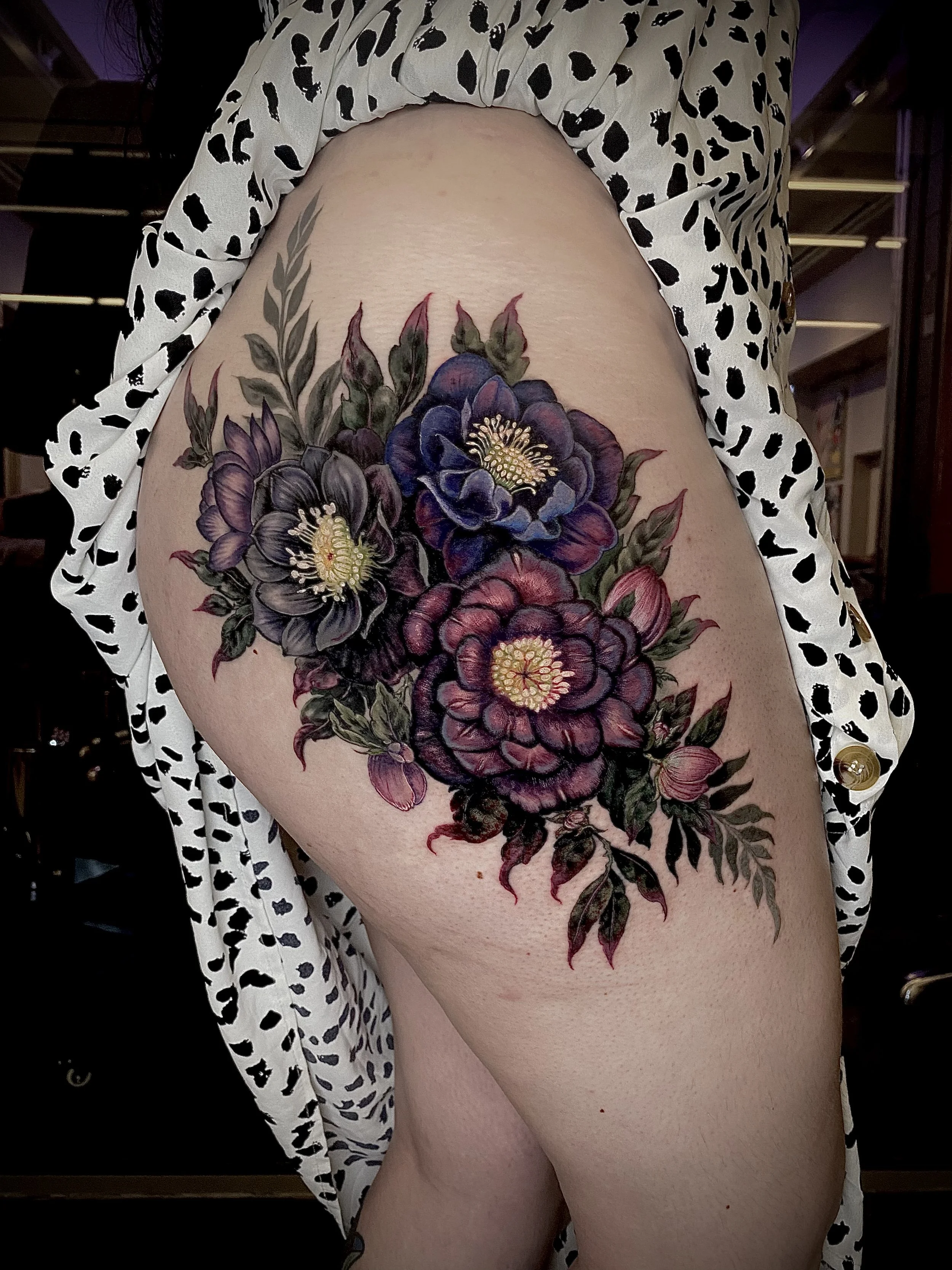 Close-up of a person's thigh with a large, colorful floral tattoo featuring purple, blue, and red flowers with green leaves. The person is wearing a black and white leopard print garment.