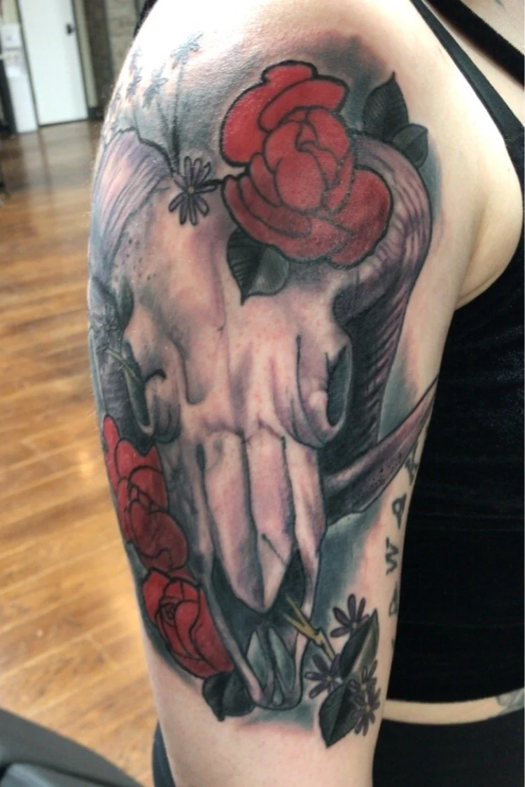 Tattoo of a skull with a large red rose on top and smaller roses and flowers around it on a person's upper arm.