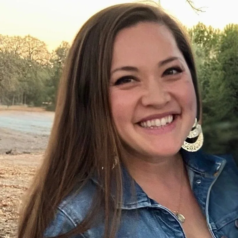 A smiling woman with long brown hair, wearing large earrings and a denim jacket, outdoors near trees during sunset.