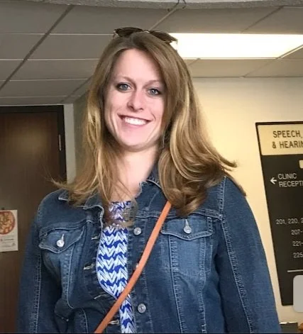 A woman smiling and posing for a photo indoors, in a casual setting, wearing a denim jacket and a patterned top.
