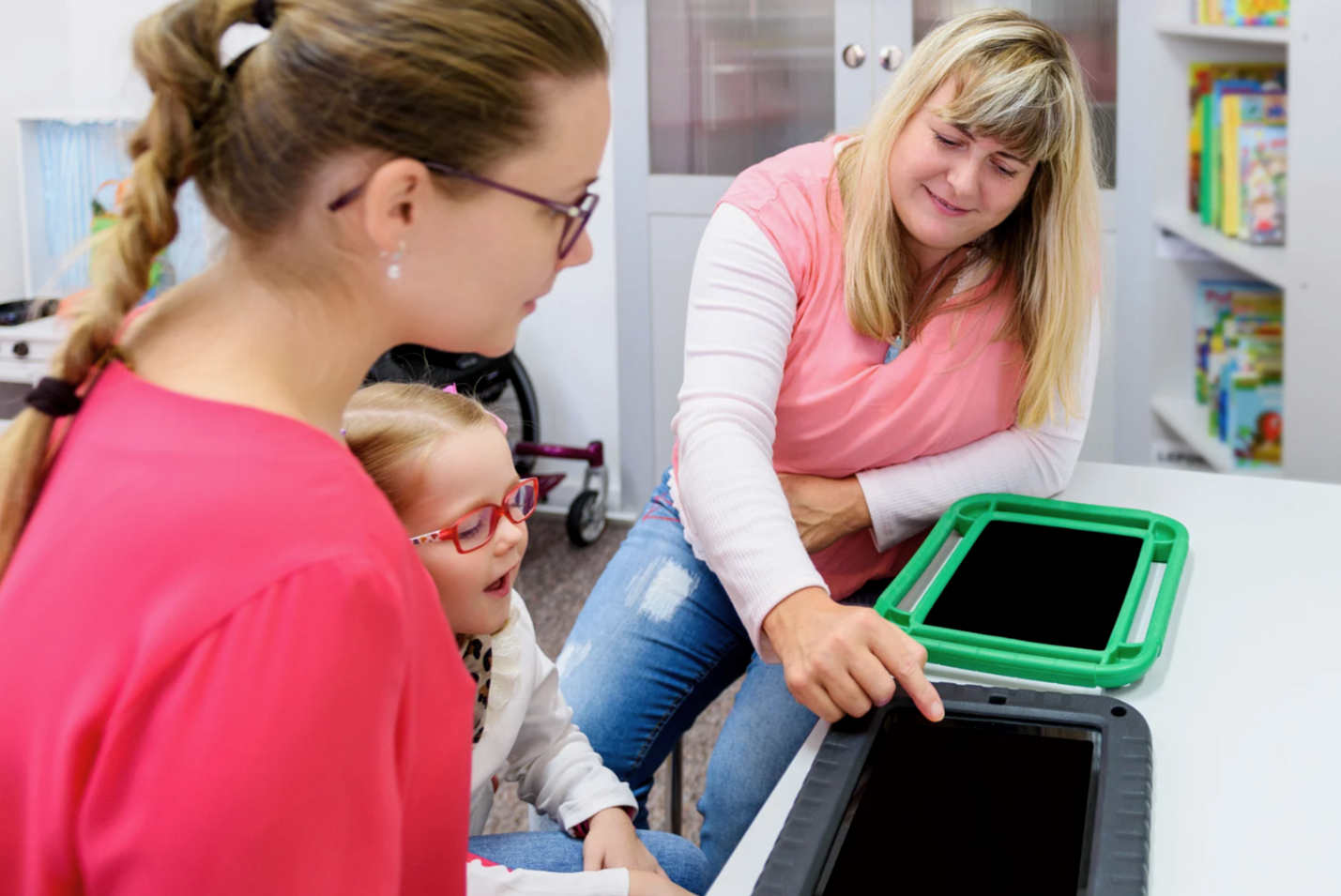 Two women and a young girl with red glasses sit at a table with two tablets, in a room with bookshelves. One woman is showing something on her tablet to the others.