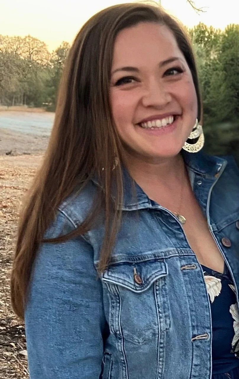 A woman with long brown hair smiling outdoors during sunset, wearing a denim jacket and large earrings.