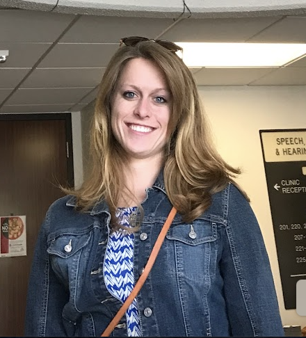 A woman with long, blond hair smiling and wearing a denim jacket, standing indoors with a sign in the background.