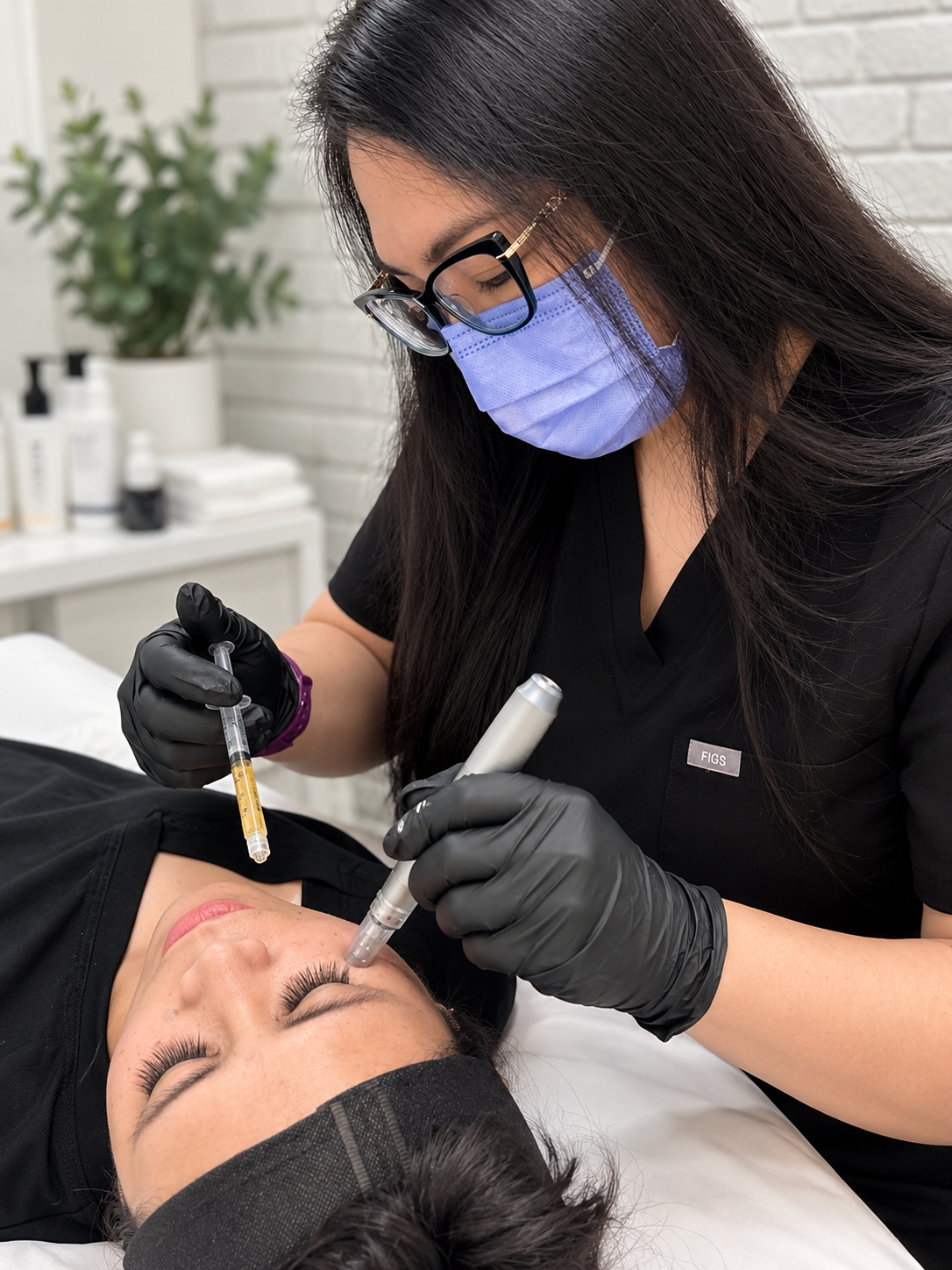 A woman in black attire, wearing glasses, a blue face mask, and black gloves, performs a cosmetic procedure on a woman lying down with her eyes closed. The setting appears to be a clinic with skincare products and a potted plant in the background.