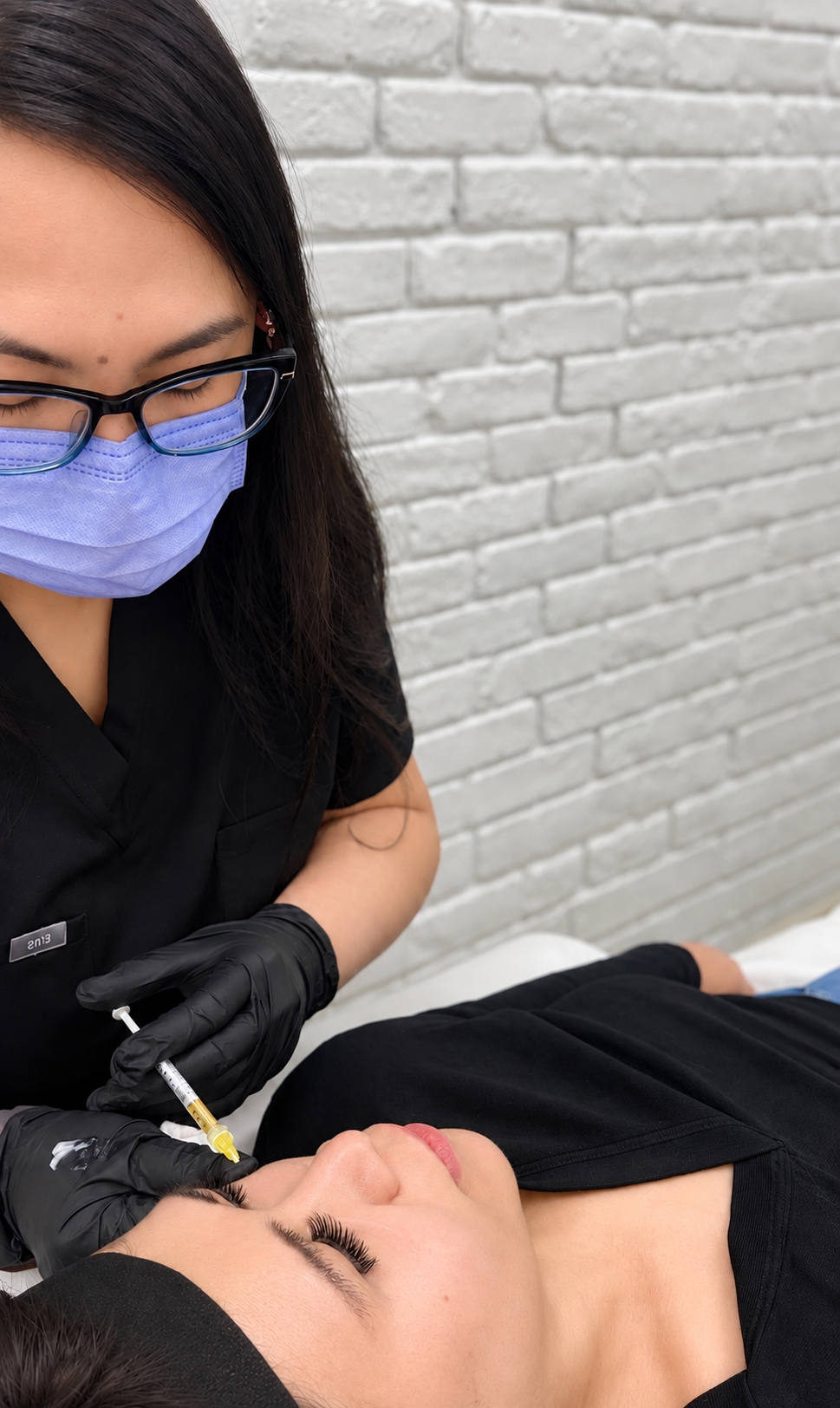A healthcare professional, wearing a blue face mask, glasses, black gloves, and black scrubs, administering a facial injection to a woman lying down with her eyes closed against a white brick wall background.