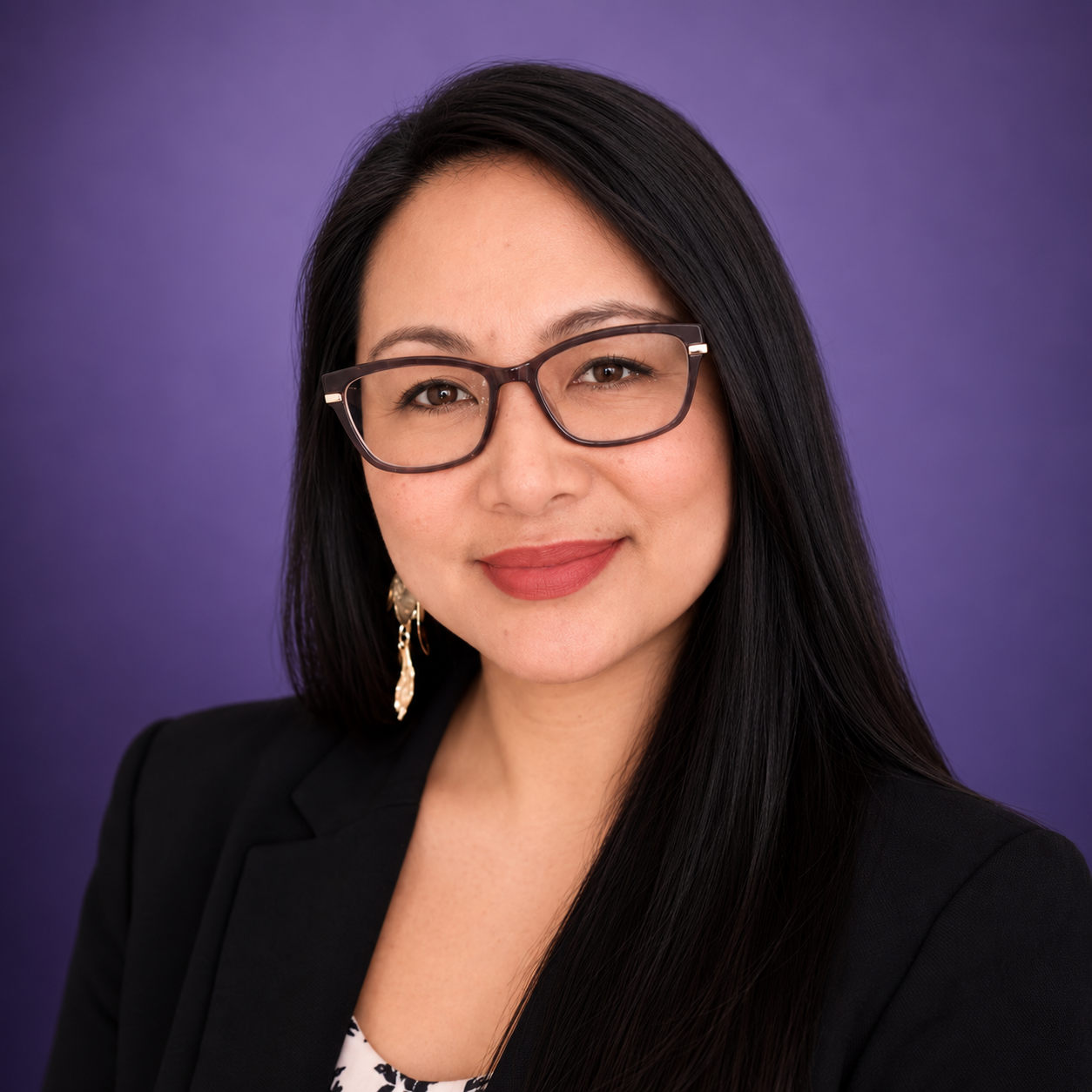 A woman with long black hair, wearing glasses, gold earrings, a black blazer, and a patterned top, smiling against a purple background.
