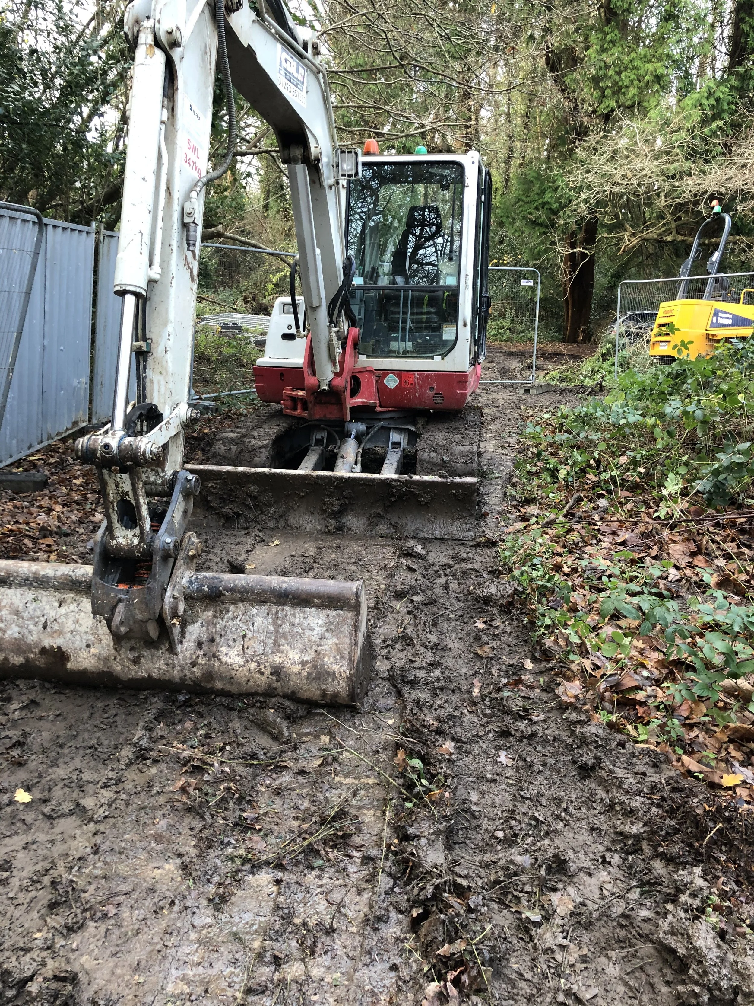 Construction site with a compact excavator digging in muddy ground surrounded by trees and fencing.