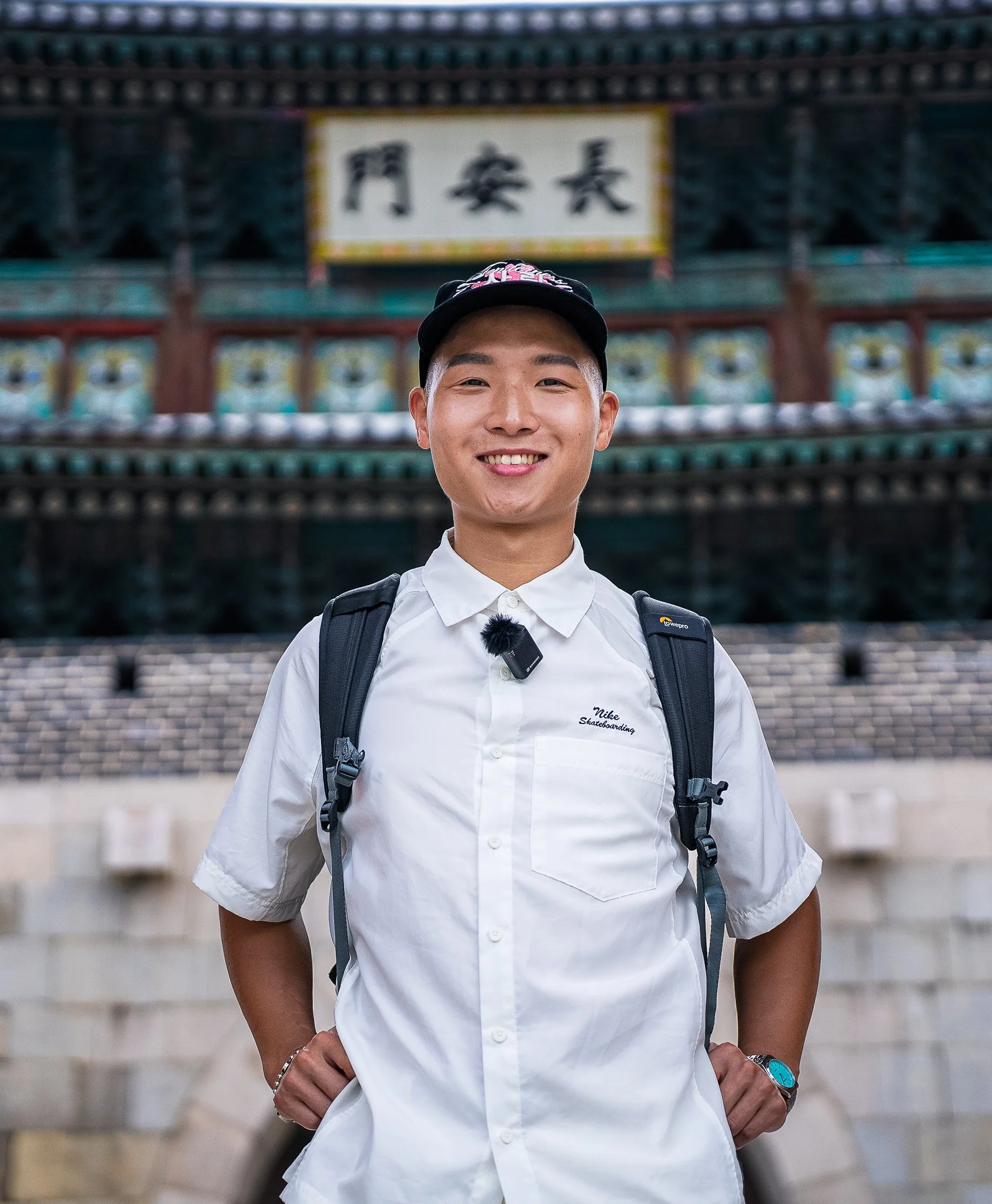 In Suwon, a young man wearing a white Nike skateboard shirt, a black cap, and a backpack, standing in front of a traditional SouthKorean building with Chinese characters on a sign, smiling at the camera.