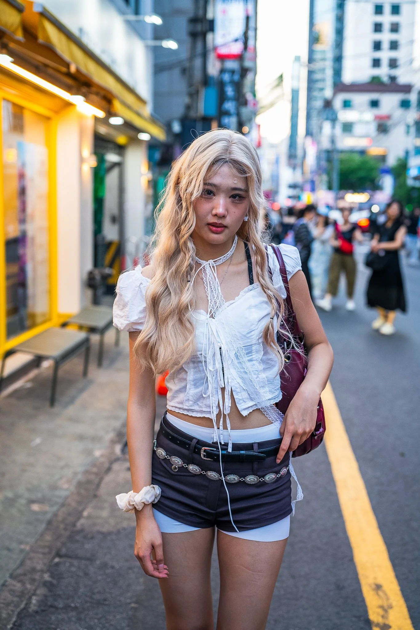 Young woman with long blonde hair and light skin walking on a busy city street in the evening, wearing a white crop top and black shorts, with a maroon bag over her shoulder.