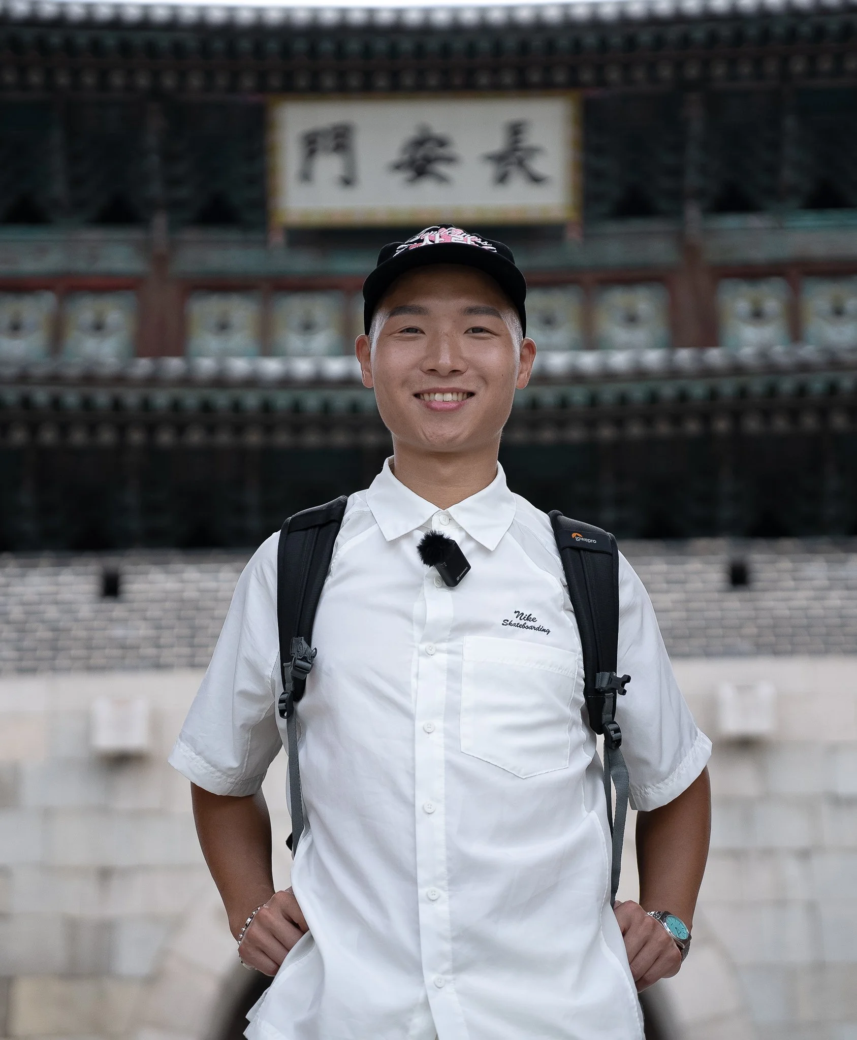 In Suwon, a young man wearing a white Nike skateboard shirt, a black cap, and a backpack, standing in front of a traditional SouthKorean building with Chinese characters on a sign, smiling at the camera.
