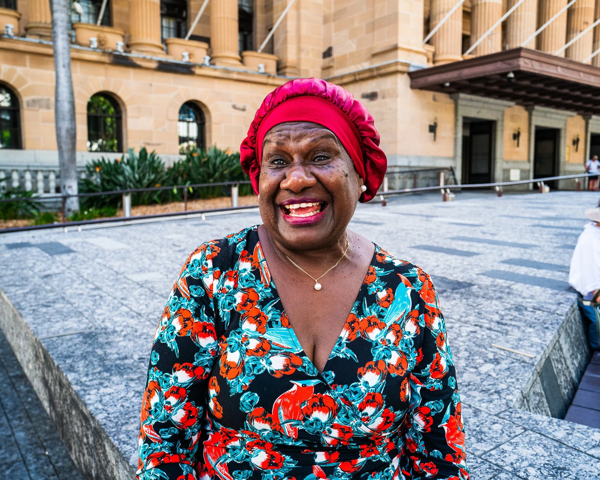 Smiling woman from Torres Strait Islands wearing a red headscarf and a floral dress outdoors in front of a historic building with columns and arched windows.