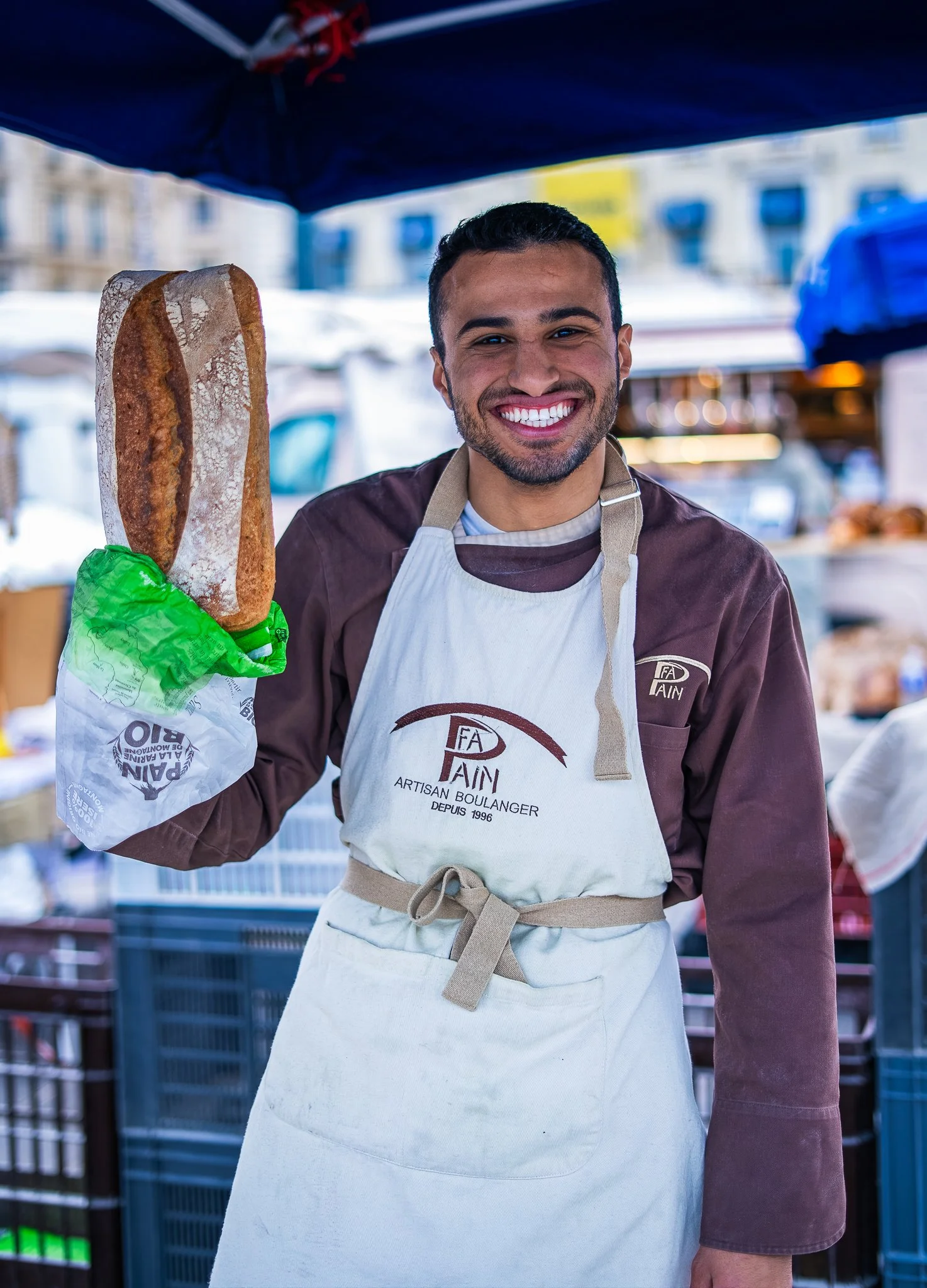 A smiling man at an outdoor market holding a large baguette in a green paper bag, wearing a apron with the logo 'FA PLAIN' and 'Artisan Boulangin Depuis 1996', with market stalls and buildings in the background.