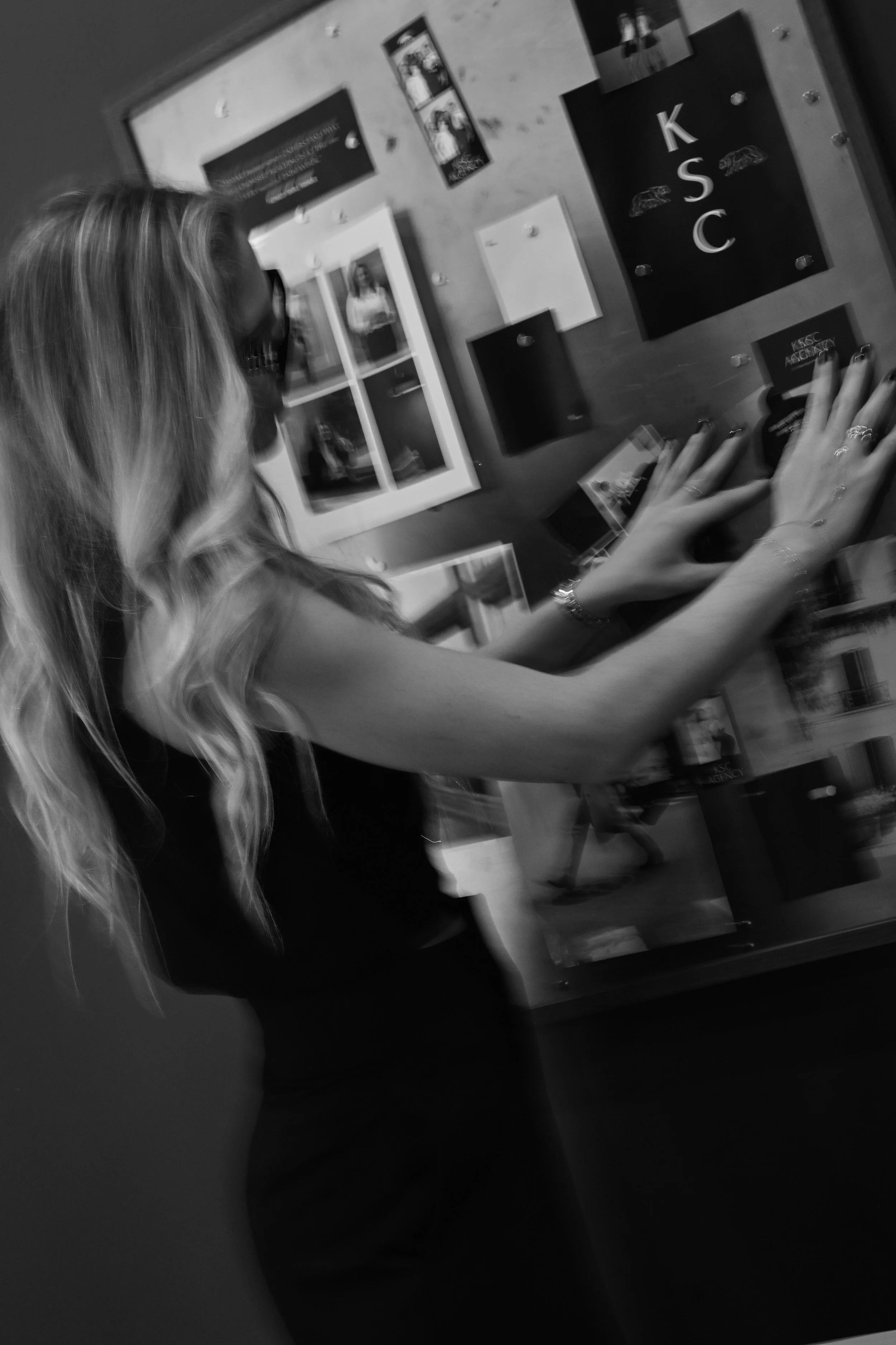 Woman with long wavy hair and glasses pointing at a blackboard with the letters K, S, C on it, in a classroom or office setting.