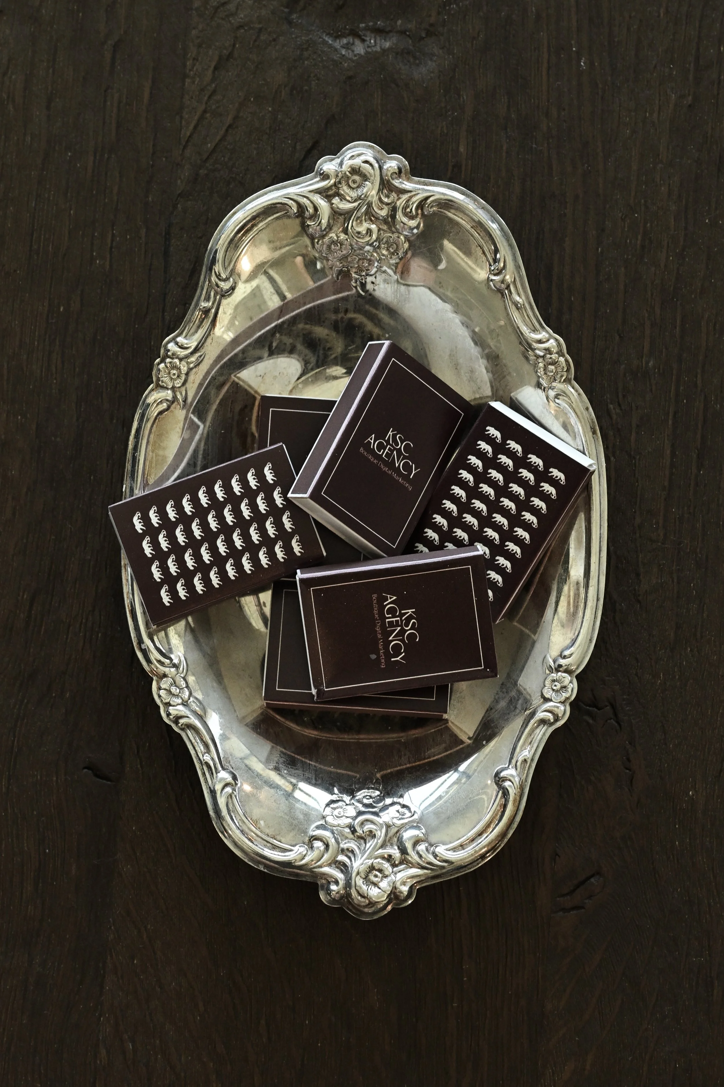 An ornate silver tray holding multiple business cards with black backgrounds and white and gold text. The cards display the text 'KSC AGENCY' and a pattern of white bears.